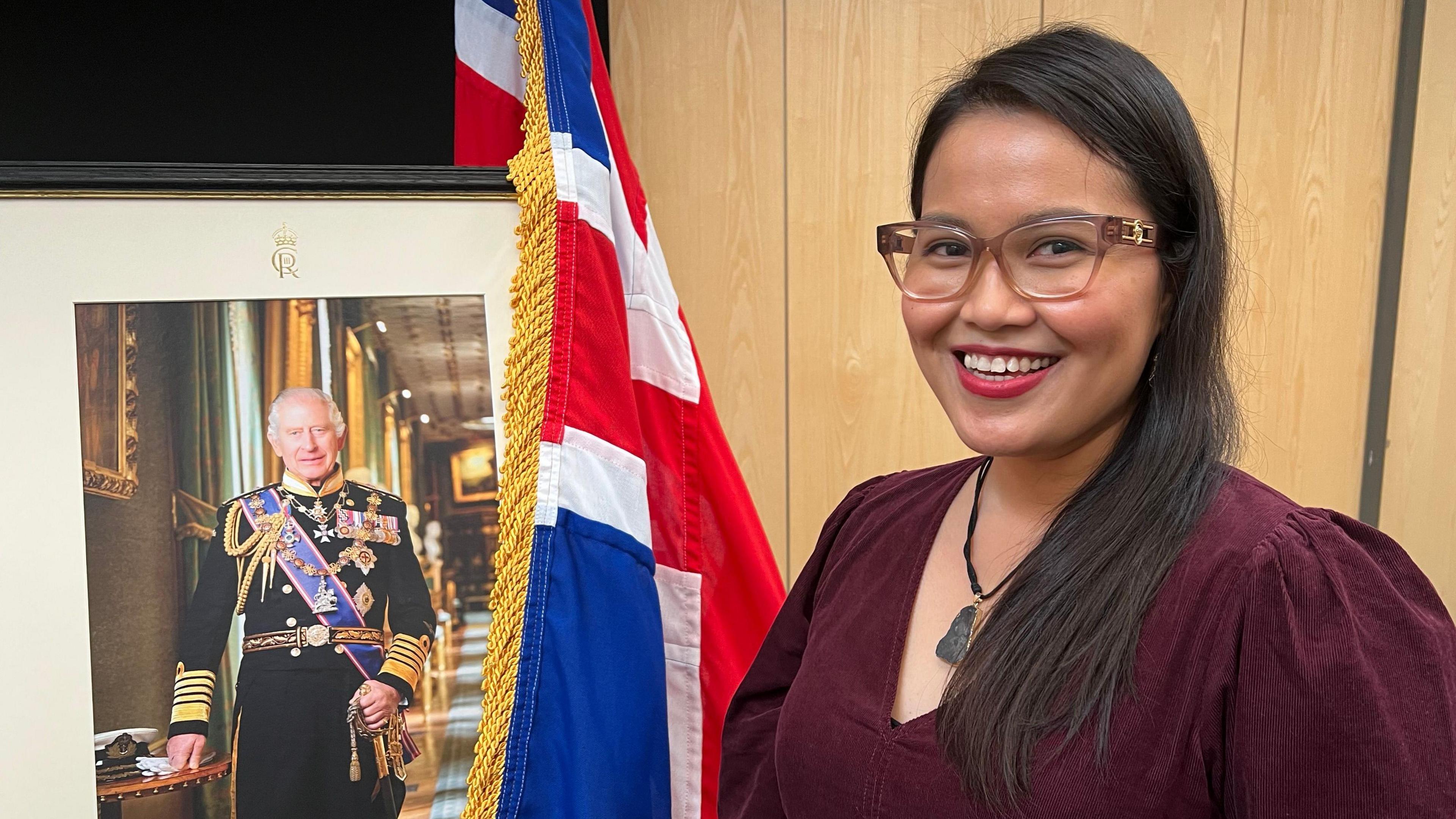 An Asian woman wearing glasses and a burgundy dress stands next to a picture of the King which is draped by a Union Jack