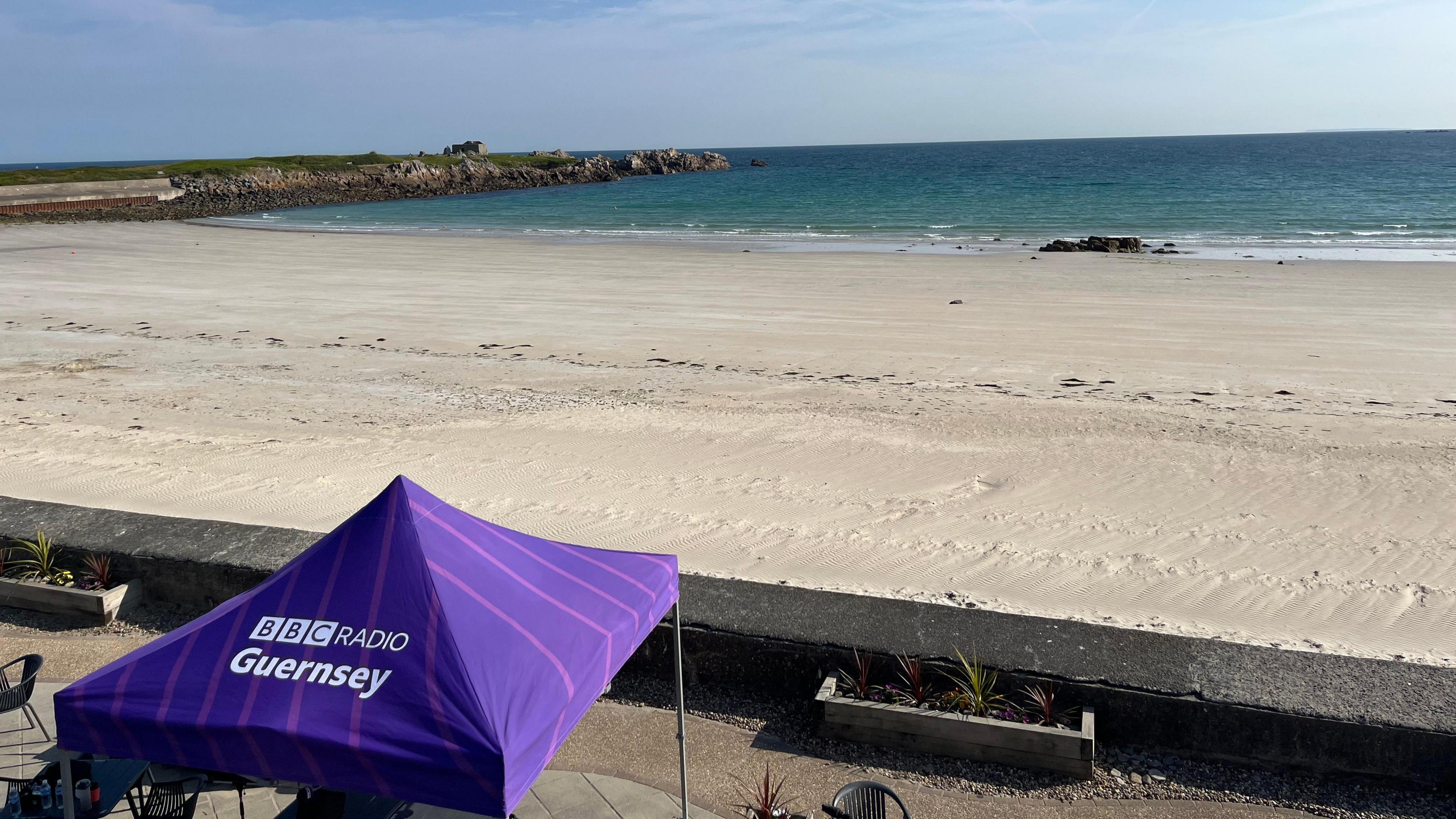 A sandy Pembroke Bay, with the tide a third of the way up the shore, with a purple BBC Radio Guernsey tent in the foreground.