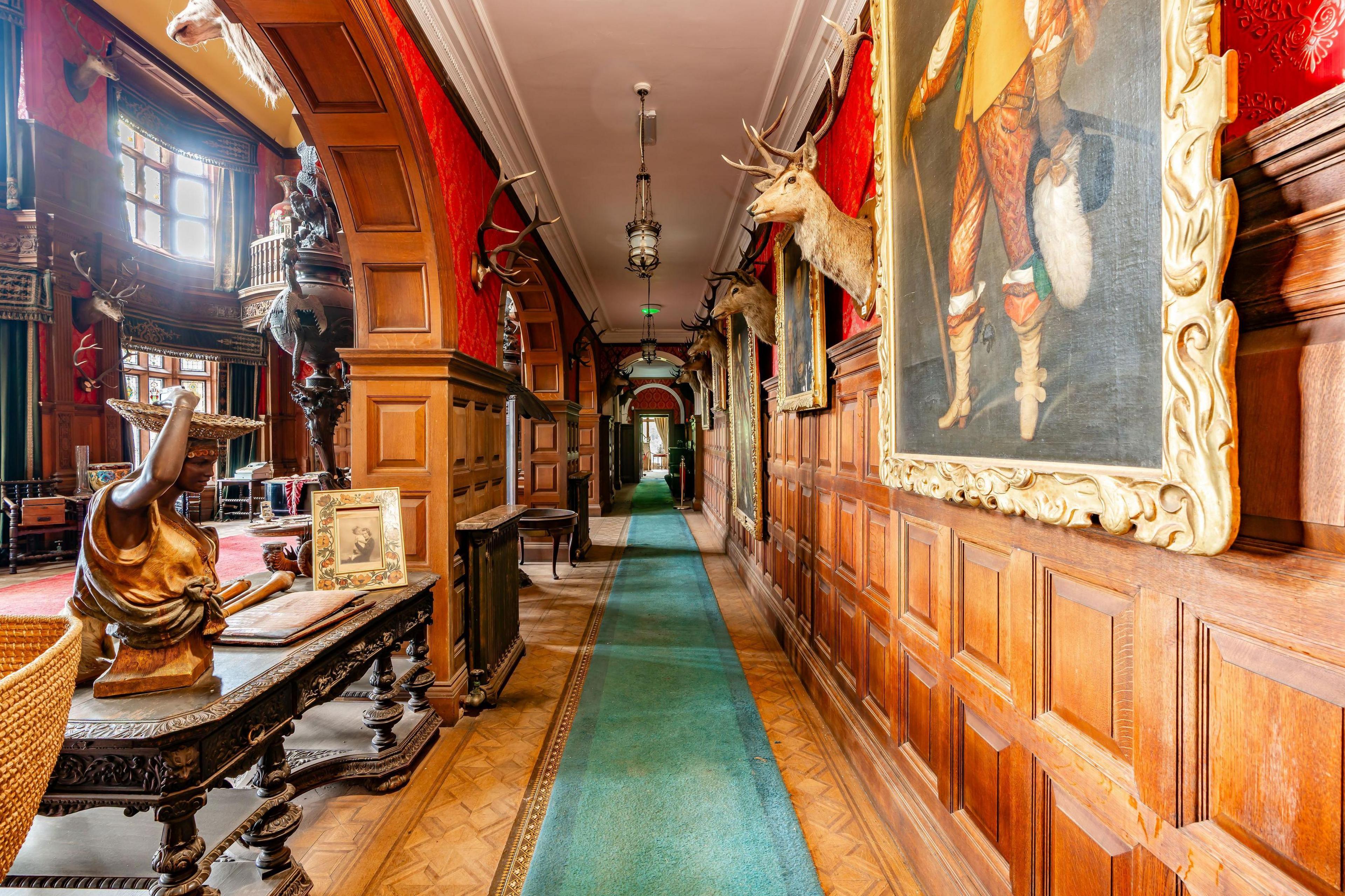 An interior view of a long corridor with wood-panel walls adorned with deer heads and paintings.