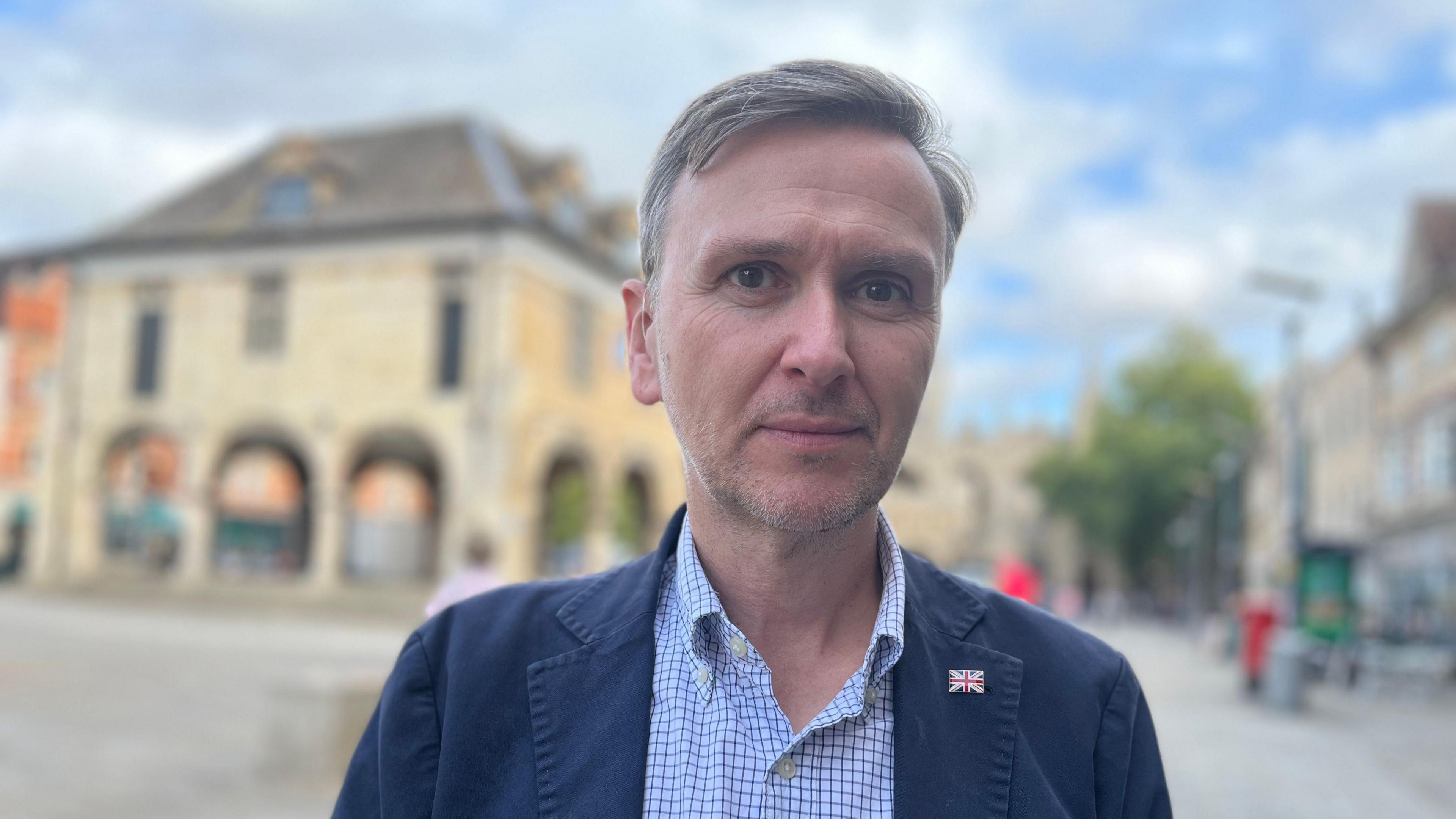 Andrew Pakes, who has short grey hair and is wearing a blue jacket over an open necked blue and white checked shirt. Behind him is Peterborough's Guildhall 