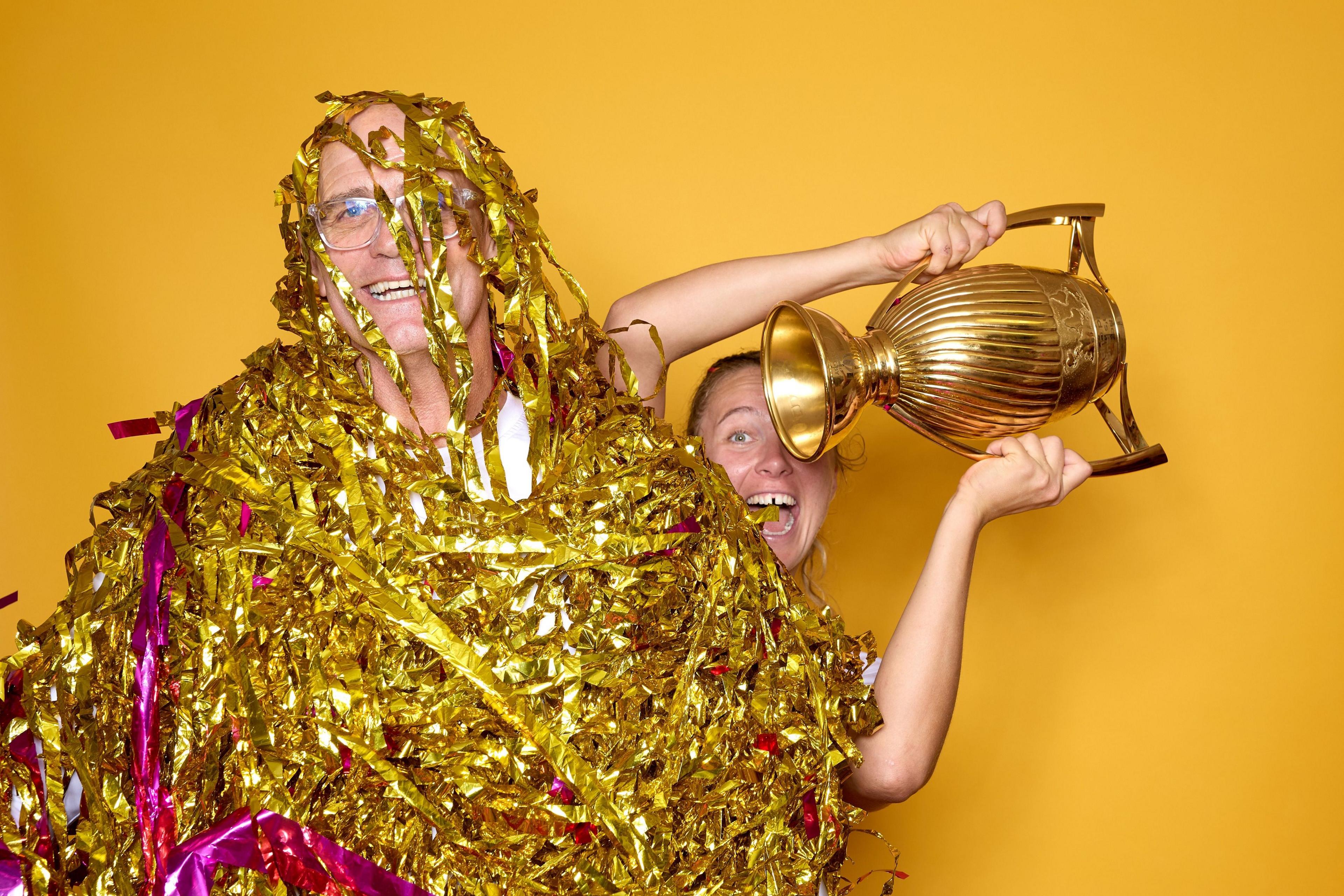 Coach John Mitchell, covered in gold and purple streamers, and captain Zoe Aldcroft, holding the trophy, pose for a portrait after England win the Women's Rugby World Cup