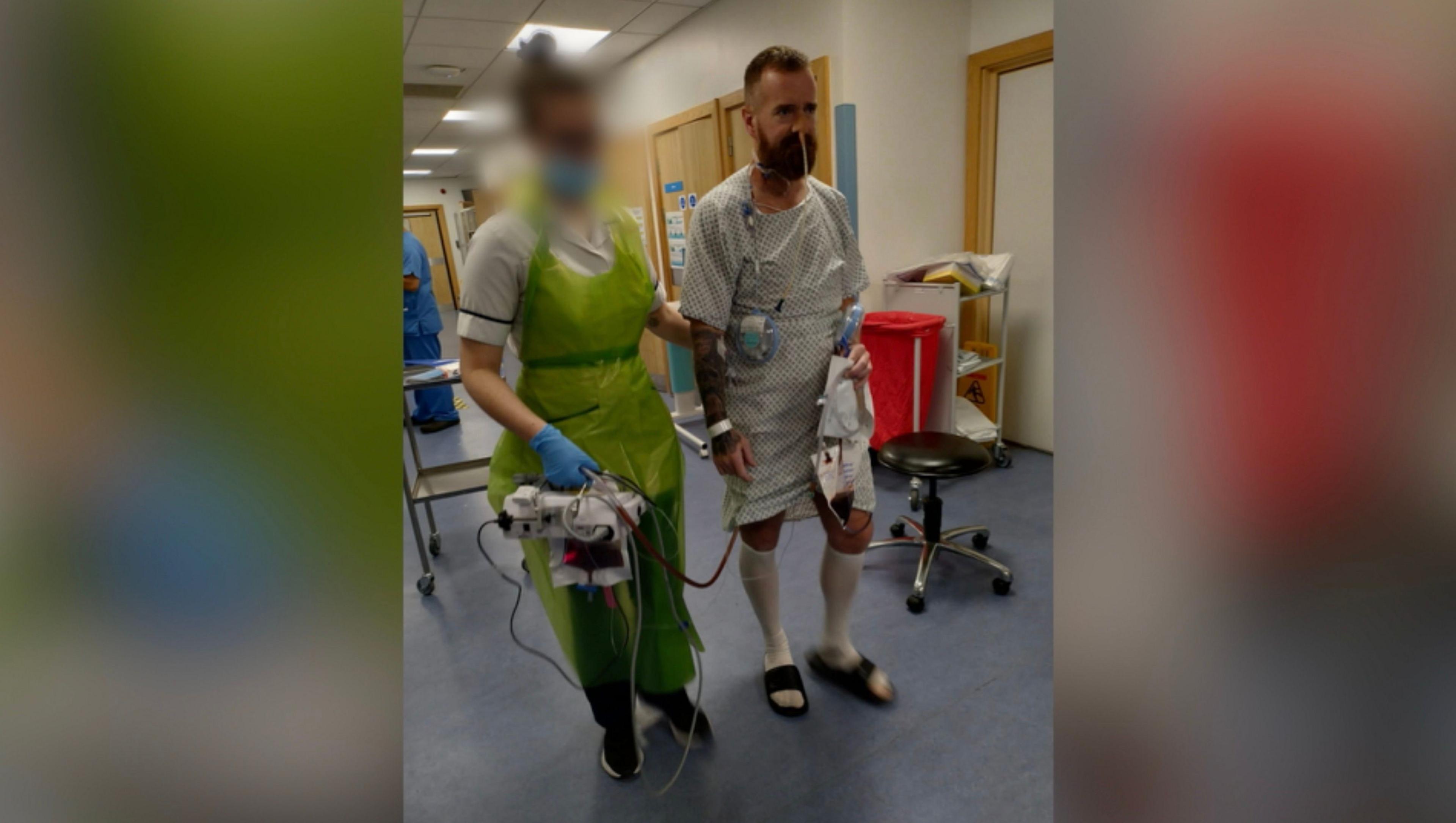 A man with a beard is wearing a hospital gown and surgical socks with sliders.  He has a tube coming out of his nose and some other tubes attached.  He is being guided by the arm by a nurse who is dressed in a plastic apron and mask.  She has glasses and her hair is tied up.