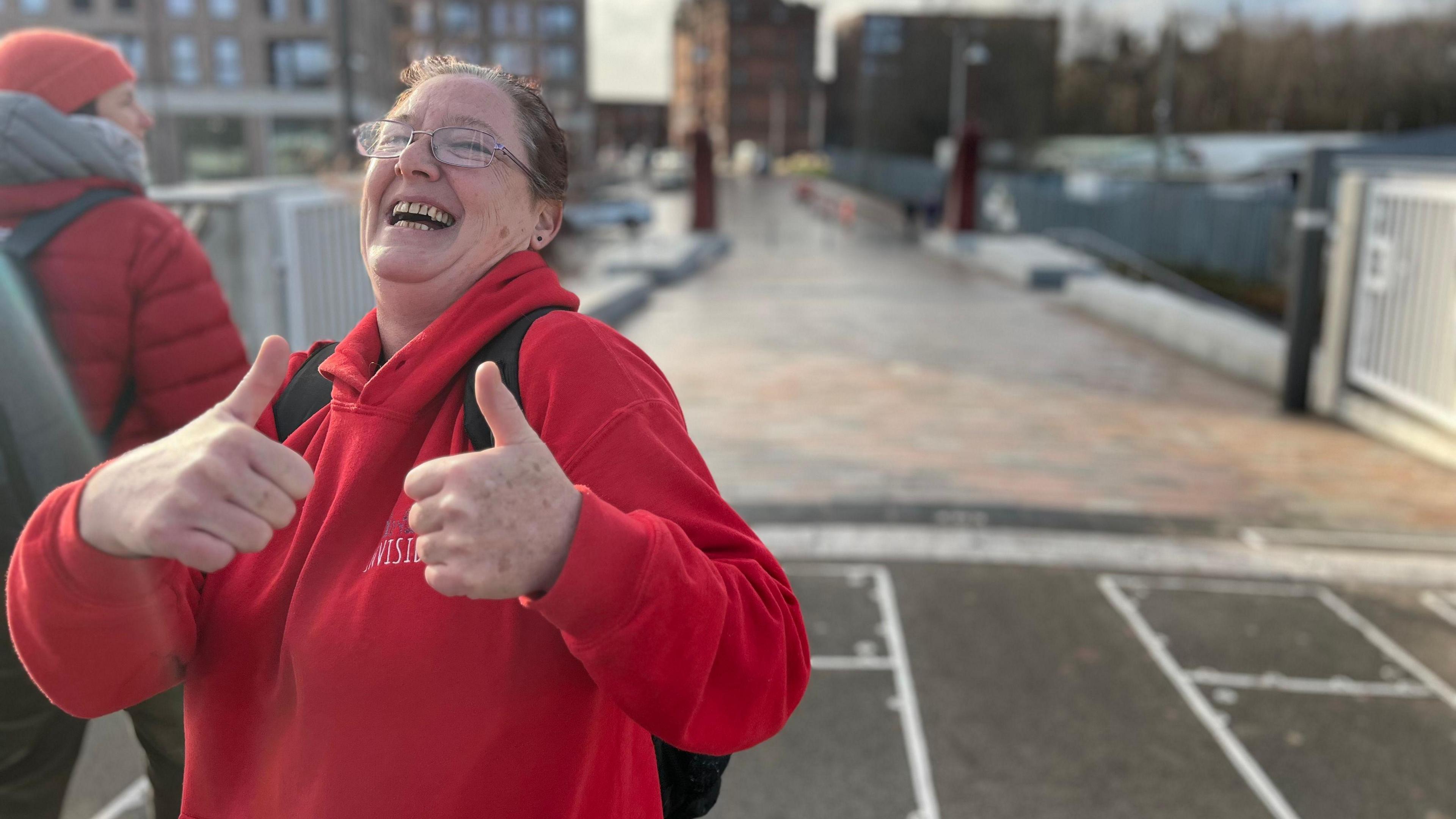 Angela McTague wearing a red hoodie. She is wearing glasses. She has her two hands raised at chest height and is giving a thumbs up pose and is laughing. She is standing on a bridge with a blurred background behind her.