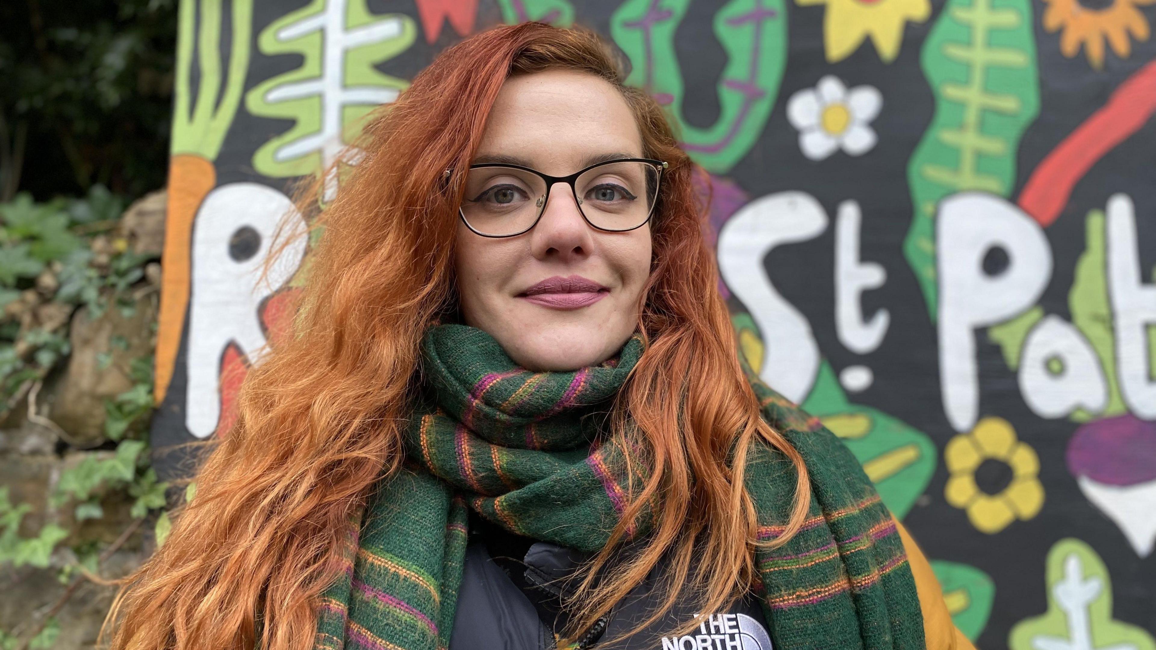 Woman stood in front of painted mural wearing a scarf and puffer jacket.