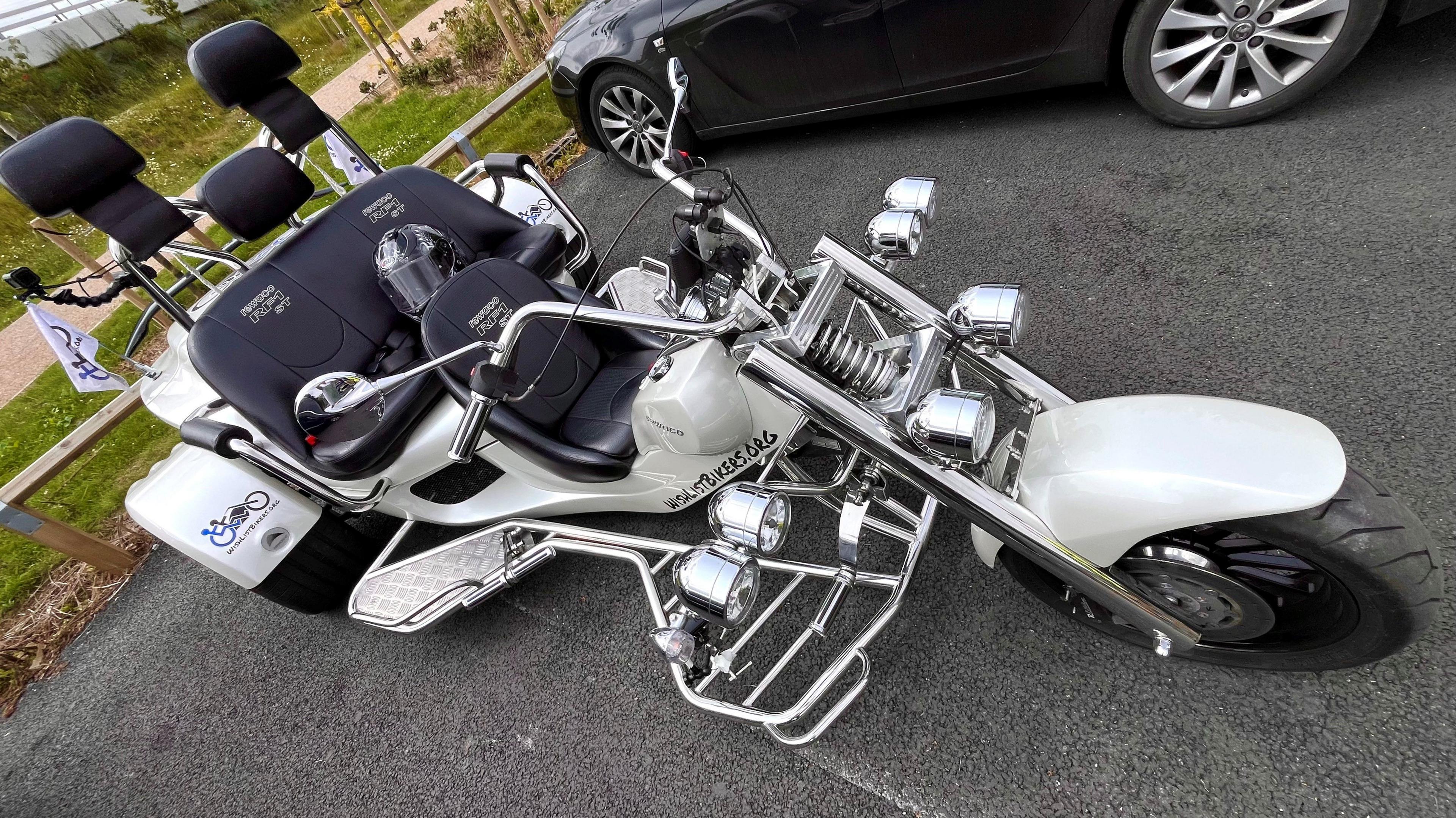 A chrome motorcycle with three wheels and wide handlebars. It has black seats. One is at the front for the rider. Two other seats are behind the rider and up higher with headrests.