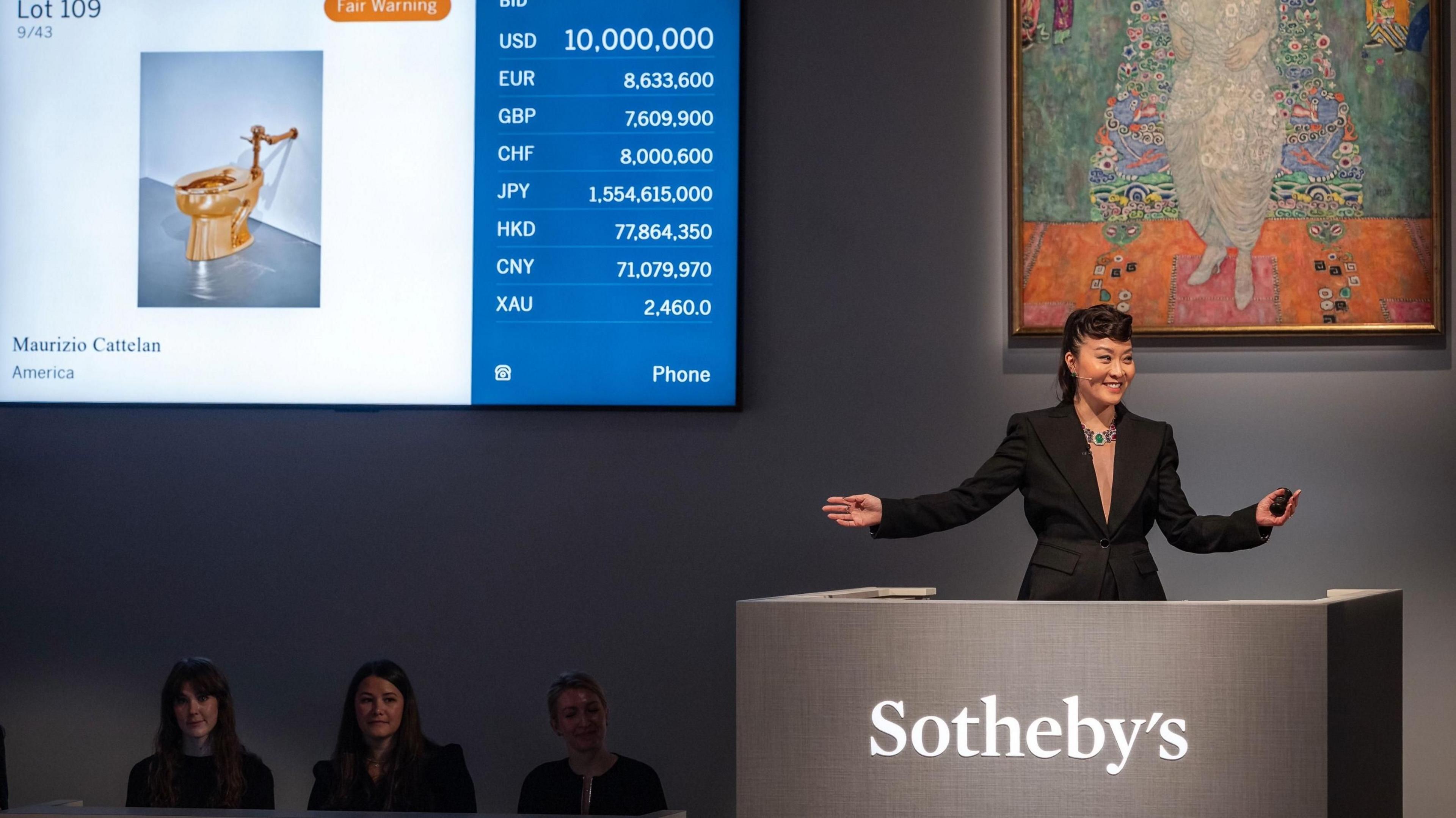 A woman in a smart black suit at a podium in the auction house. A picture of the gold toilet is on a screen. A Gustav Klimt artwork is also on show.