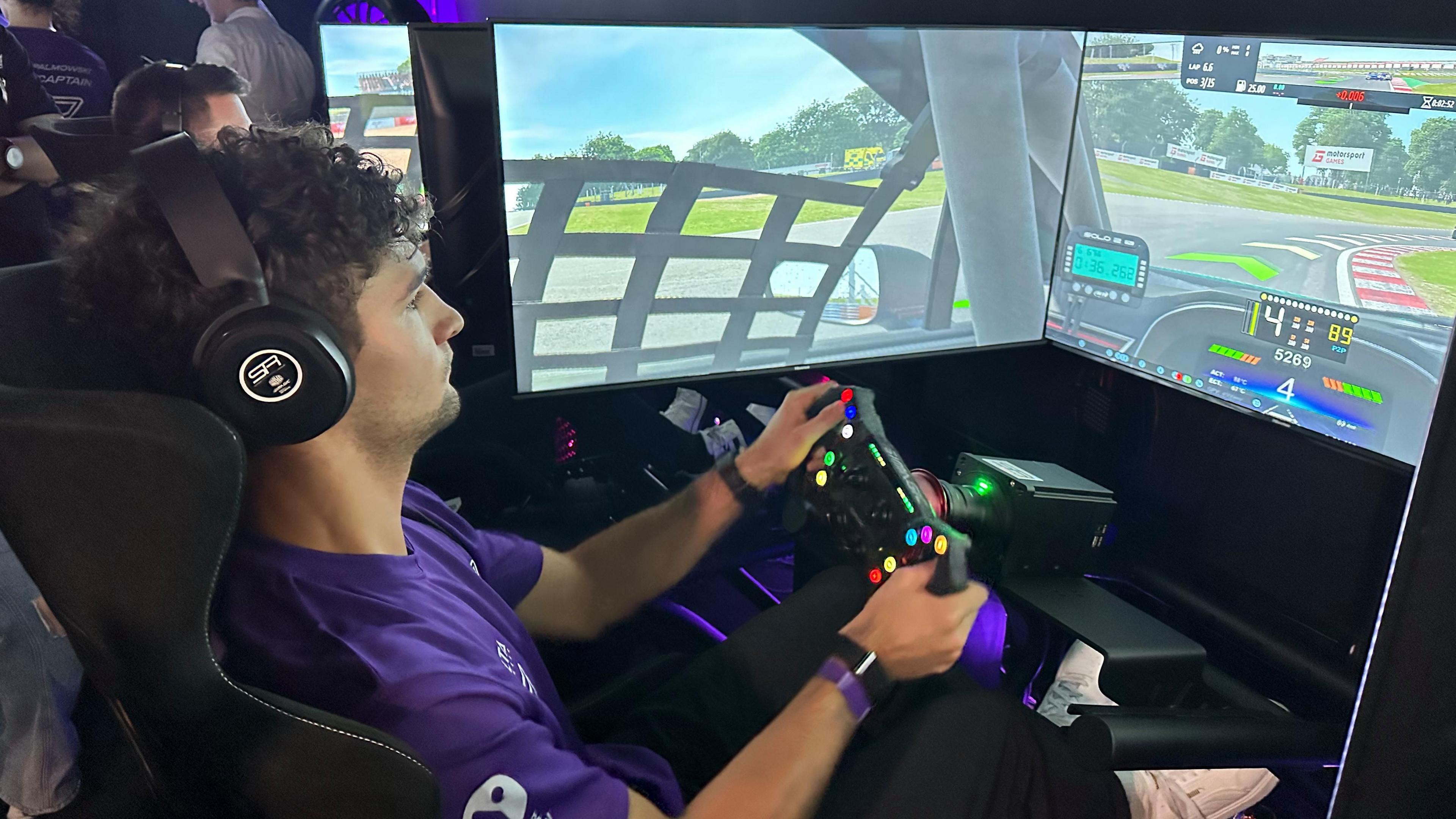 Jak Crawford, a man in his 20s with dark brown curly hair on a racing simulator. There are screens surrounding him displaying inside a race car on track.