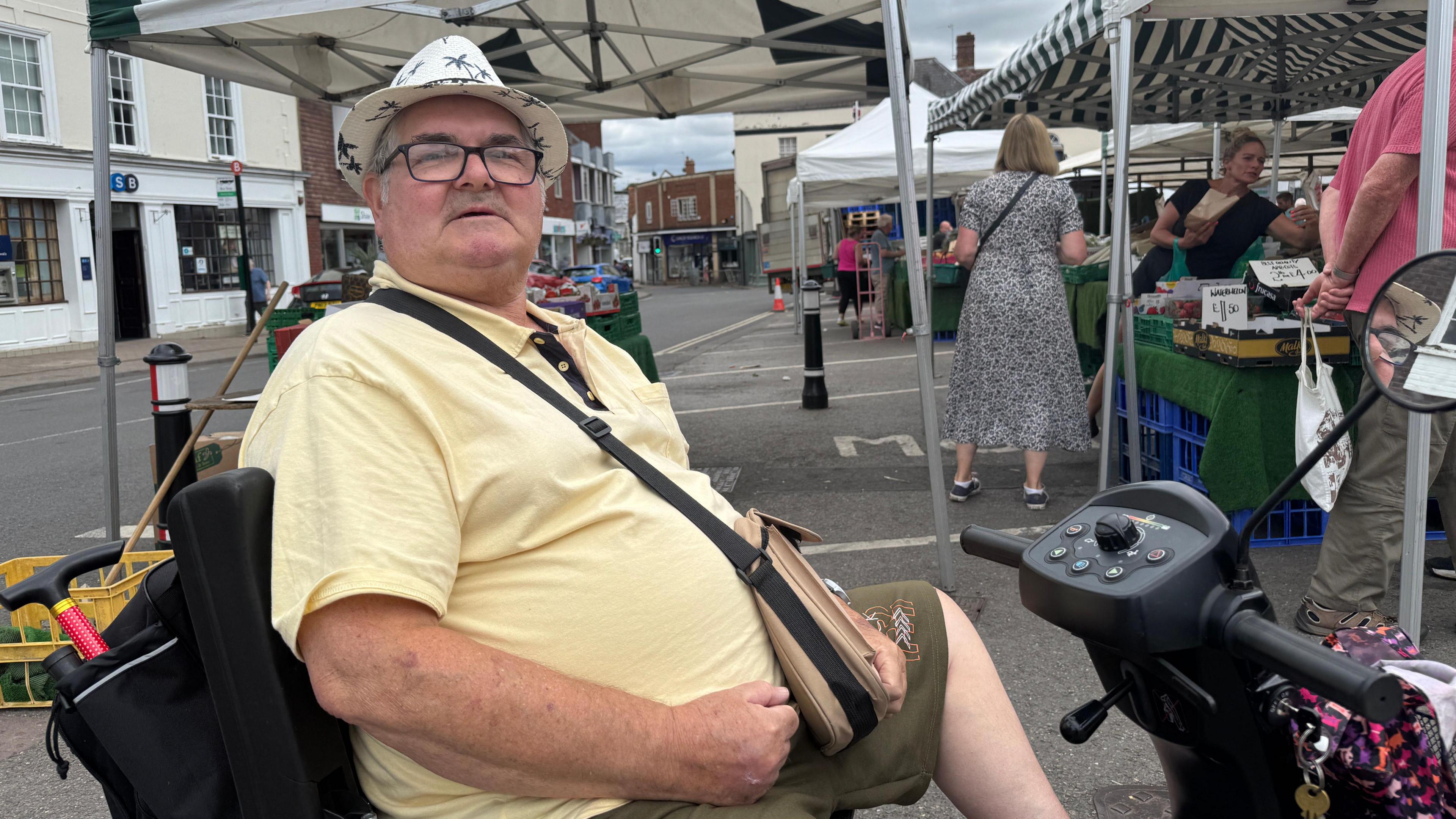 Gene Hicks sits on his mobility scooter and looks to the right towards the camera. He is wearing a yellow polo shirt and a pork pie hat with the work 'Lanzarote' on it. He has a satchel slung over one of his shoulders. The street market is visible with traders and shoppers in the background
