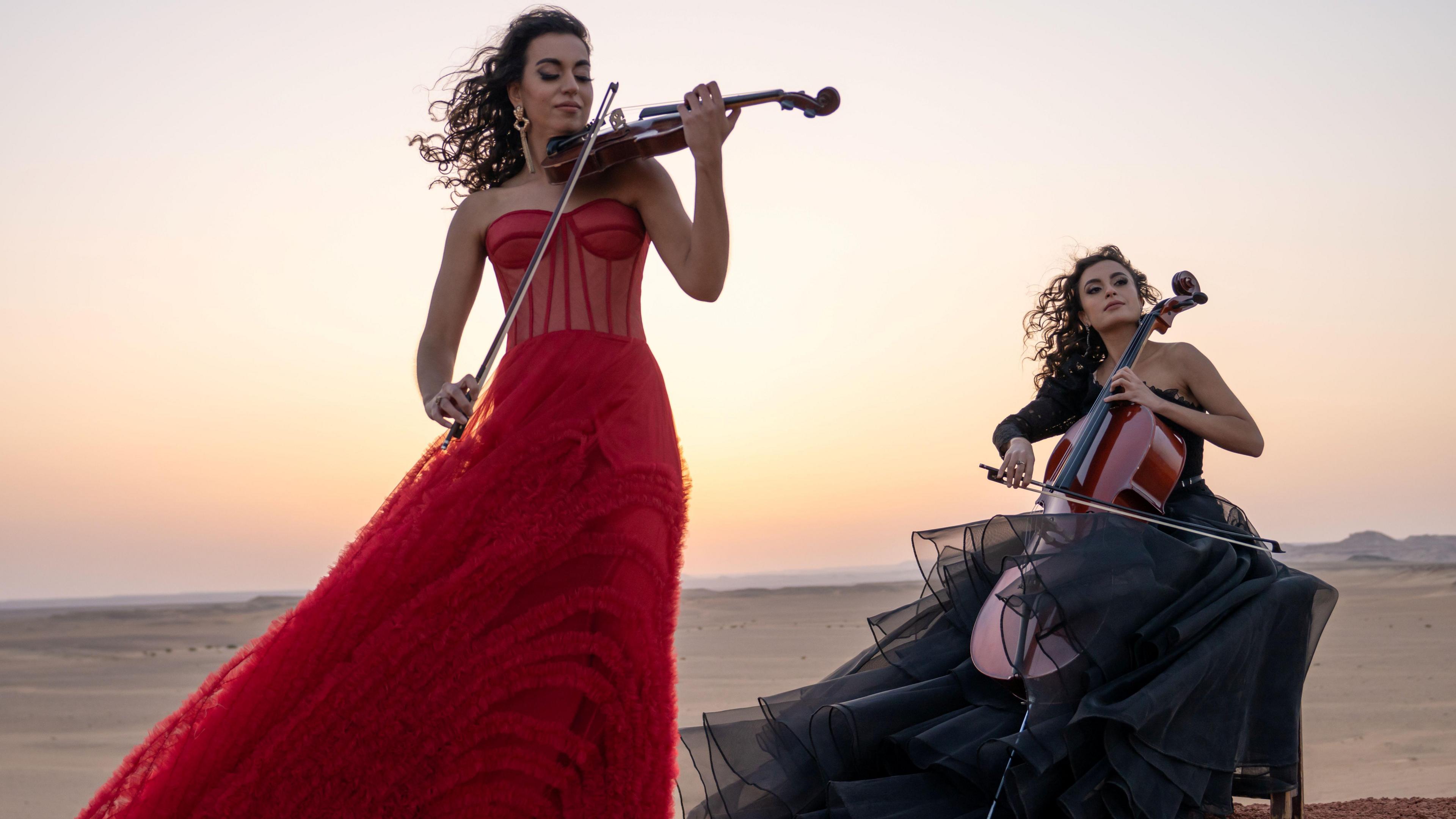 Two women outdoors in the desert, playing the violin and the cello. One woman is standing up, wearing a red dress, the other is sitting down and wearing a black dress.