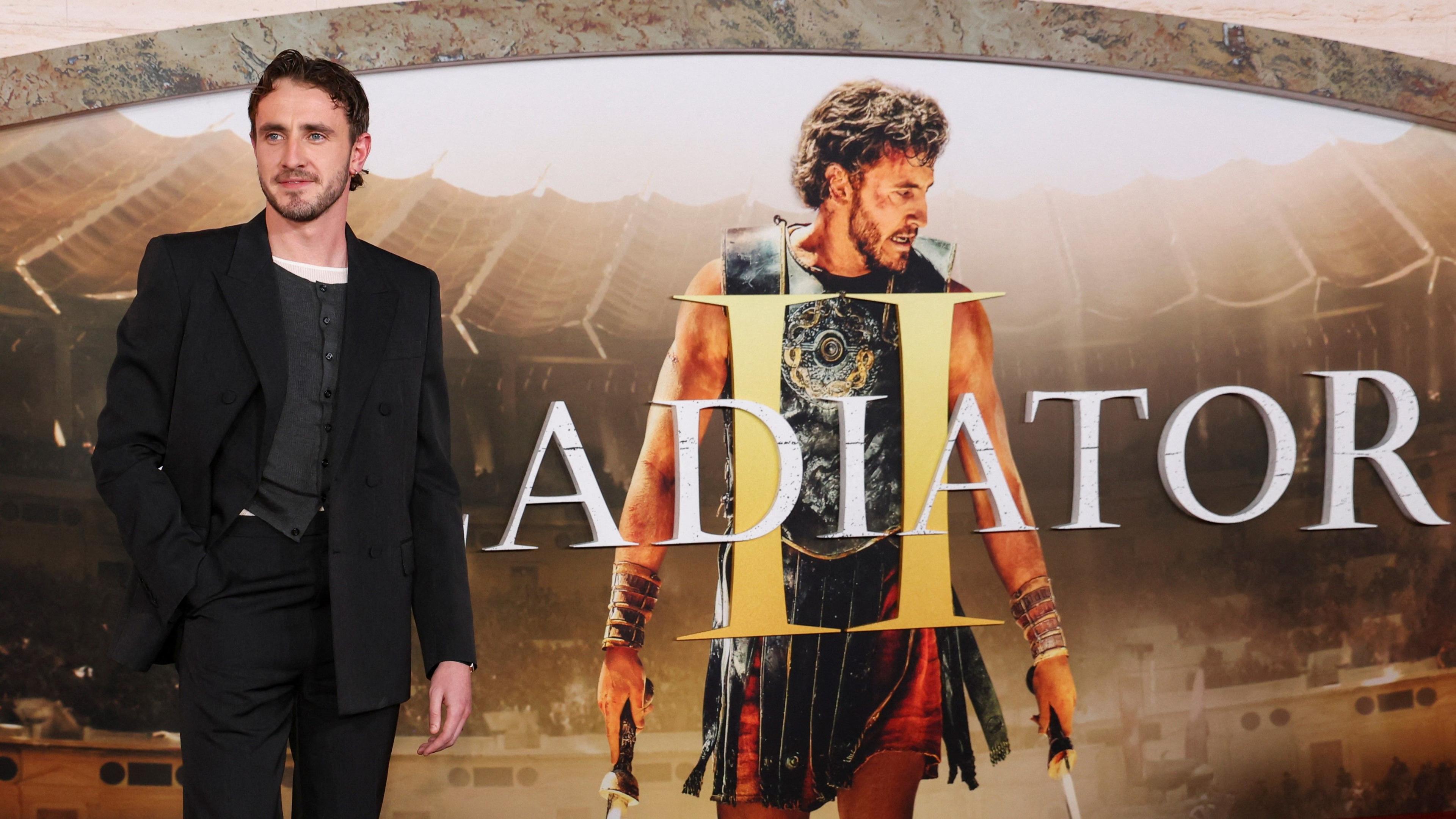 Paul Mescal, dressed in a black suit, attends a premiere for the film 'Gladiator II', standing in front of a poster with the name of the film and a picture of himself, dressed in ancient Roman costume, at the TCL Chinese Theatre in Los Angeles, California, U.S. on 18 November