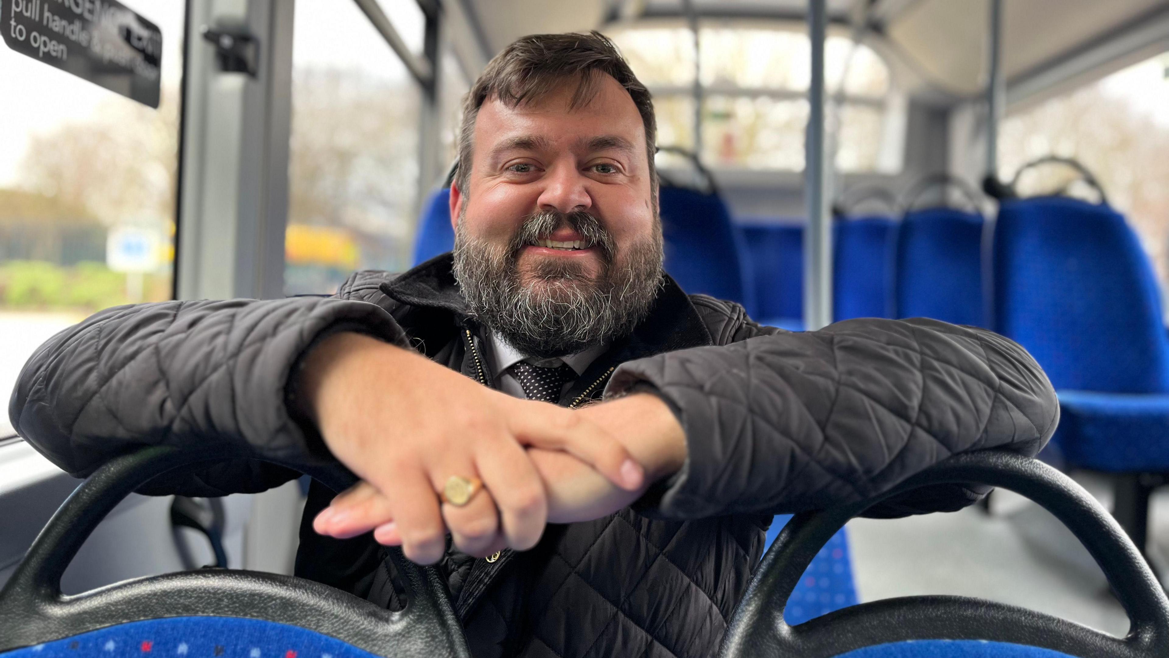 Andrew Sherrington inside a bus leaning on the seats in front and looking into the camera. He has a beard and is wearing a black quilted jacket.