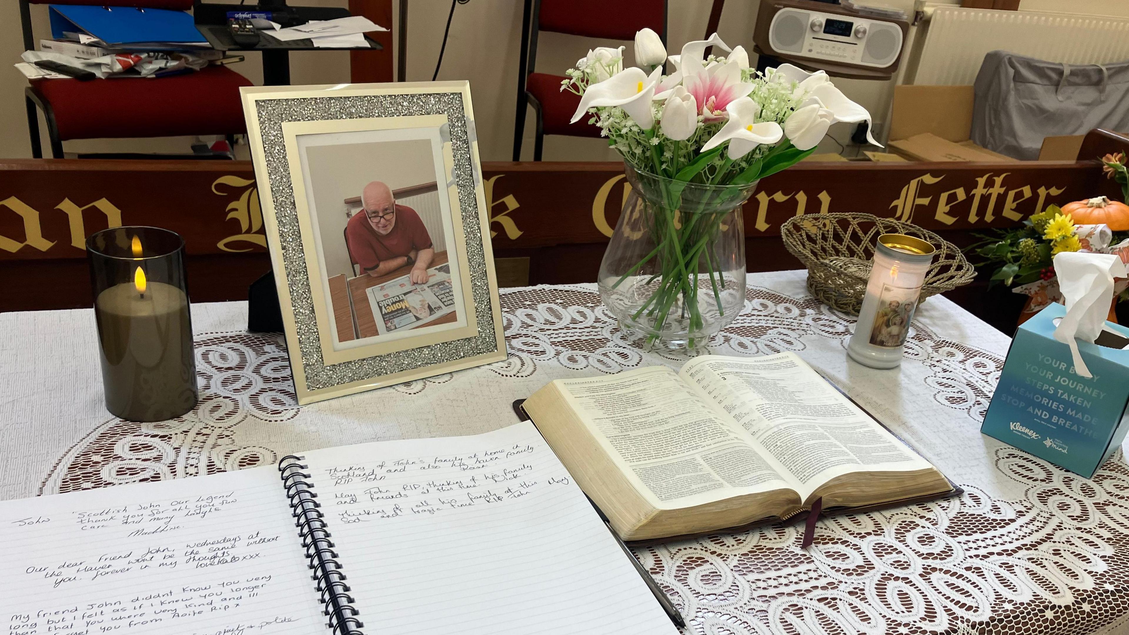 A table with two books open. A picture frame with an image of a man wearing a read top reading a newspaper. There are candles and flowers on the table.