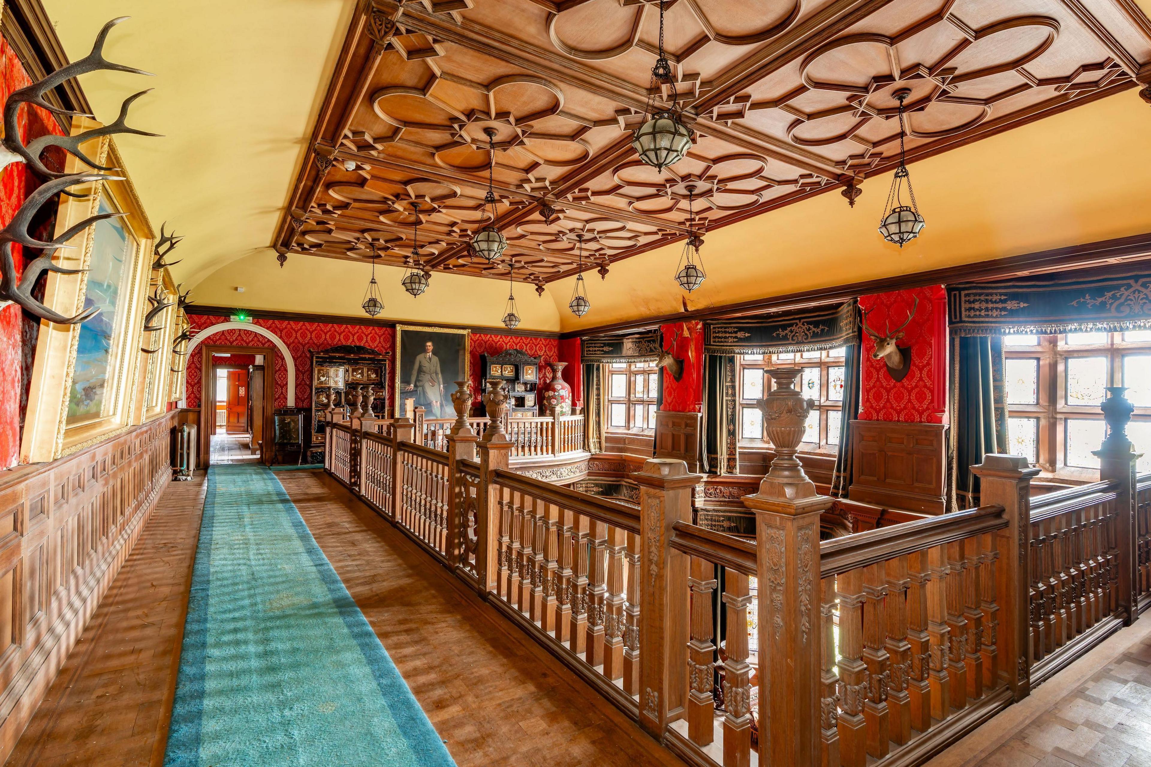 An interior view of Kinloch Castle showing an upper floor with carved, wooden banisters and a long pale blue rug. Deer heads and paintings adorn the walls. 