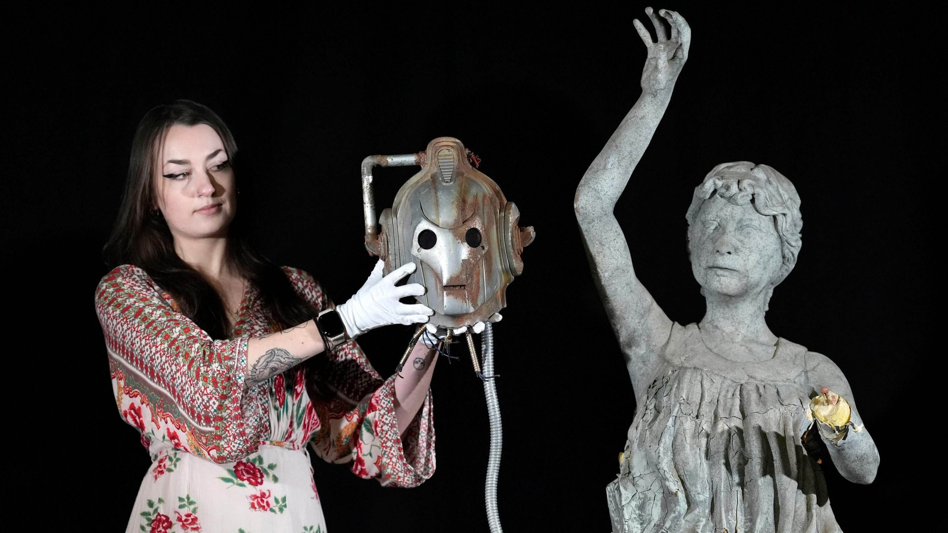 A woman is holding a decapitated Cyberman head next to a weeping Angel statue which is missing wins and a left hand.