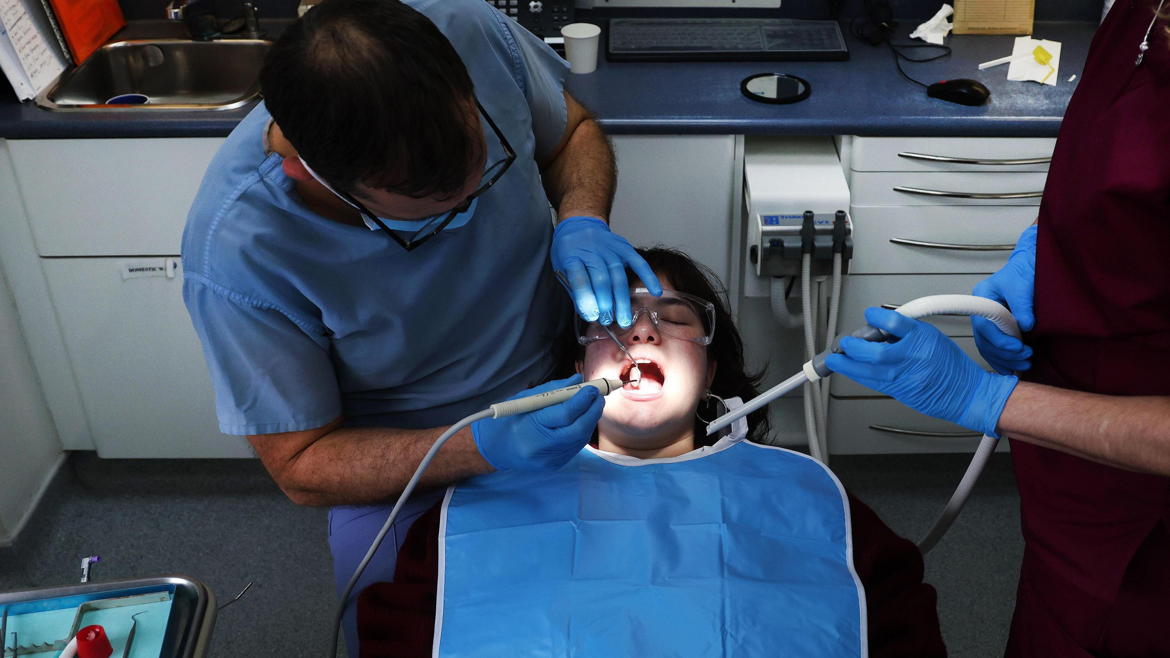 A man wearing surgical gloves and using dental equipment gives treatment to a lady with an assistant present.