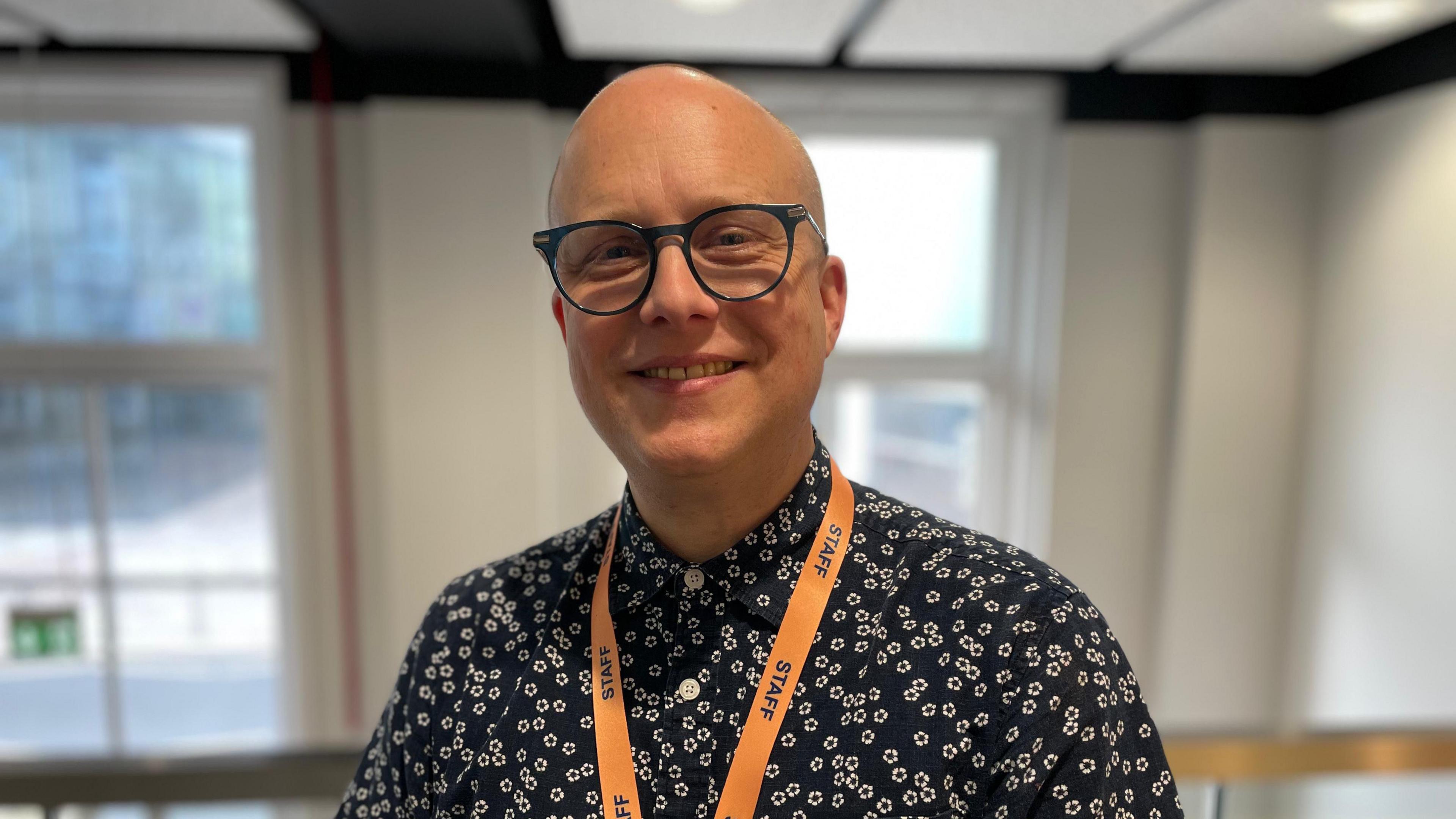 Bradford Arts Centre CEO Alex Croft is pictured. He is bald, wearing blue glasses. He wears a navy blue shirt with small white flowers on it, and has an orange lanyard around his neck.