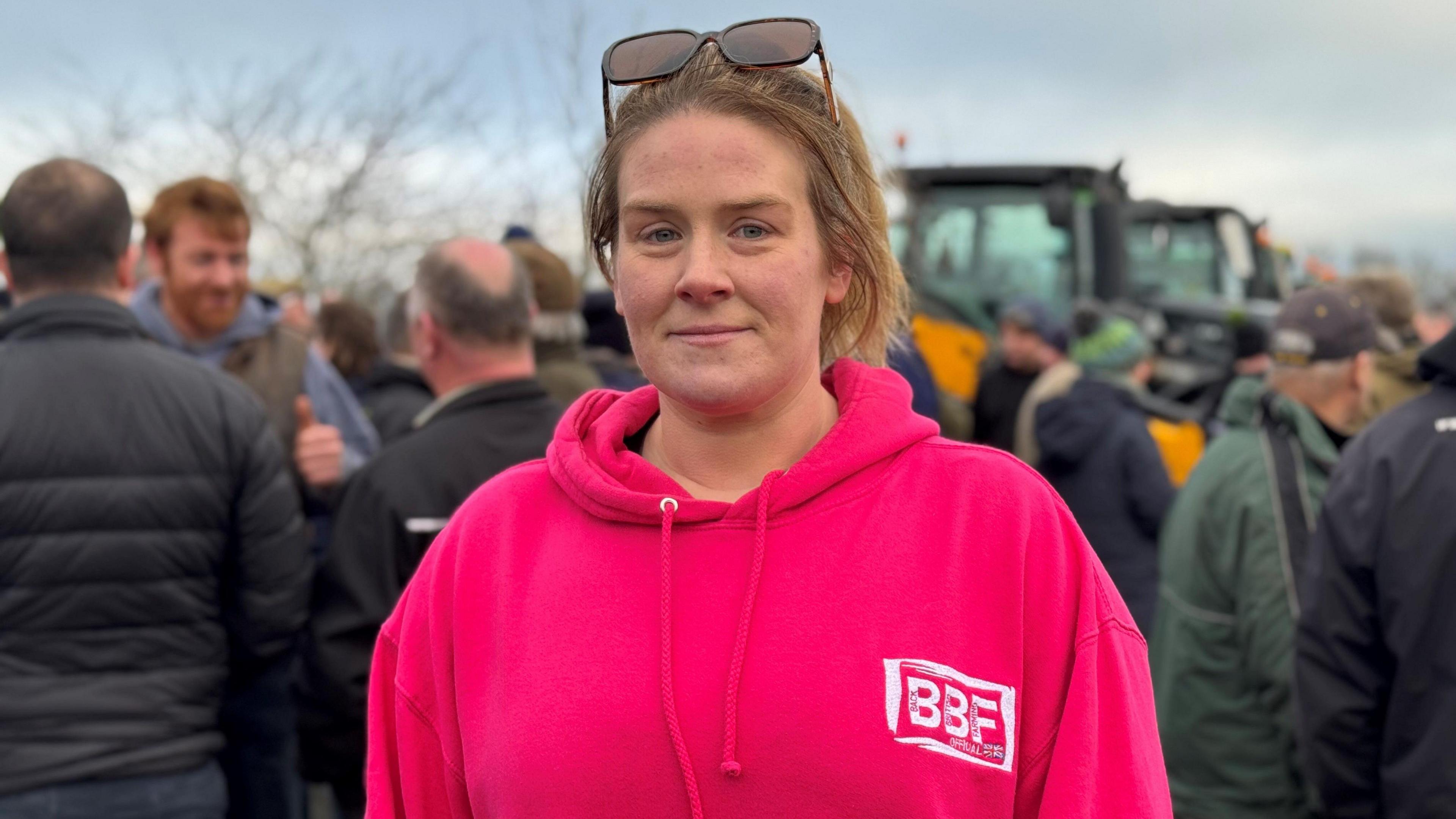 Rachel Fife is standing with tractors behind her. She's wearing a bright pink top and has blonde hair with glasses pushed onto her head 