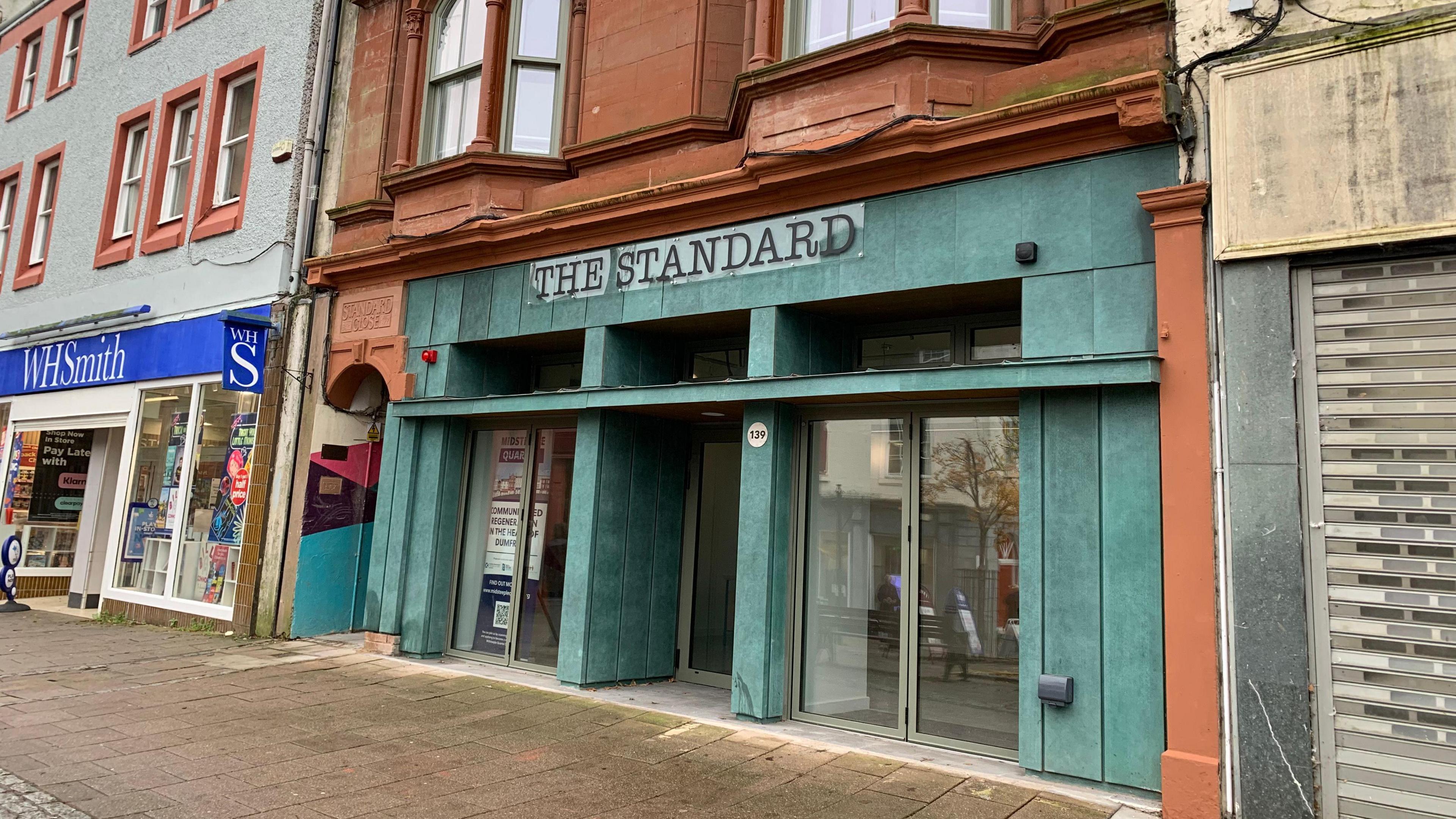 A green-fronted town centre property with sandstone surrounds sits between one empty building and a WH Smith store