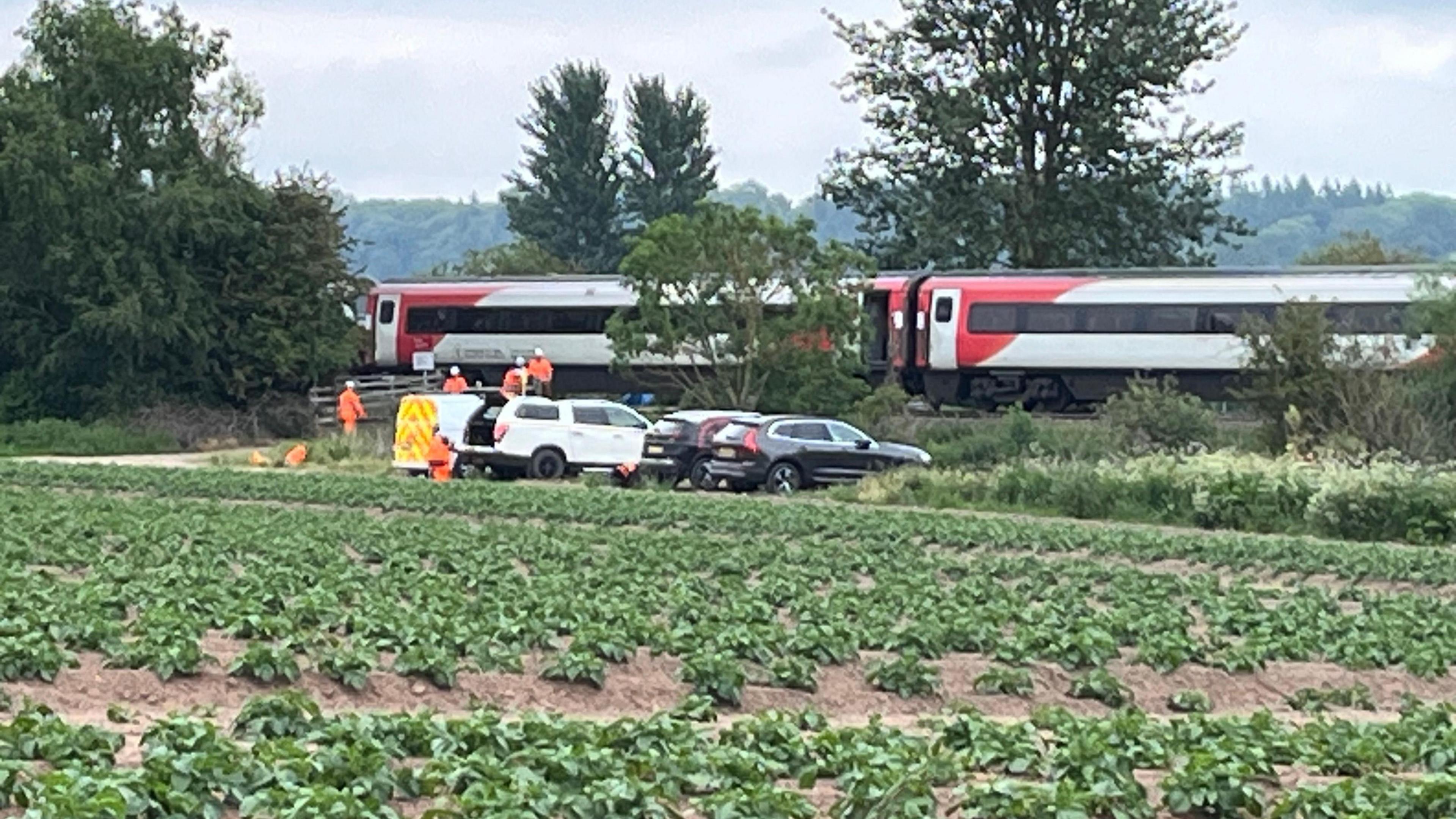 A train has come to a halt after hitting a tractor and trailer. A field can be seen in the foreground with emergency and other vehicles part in front of the train.