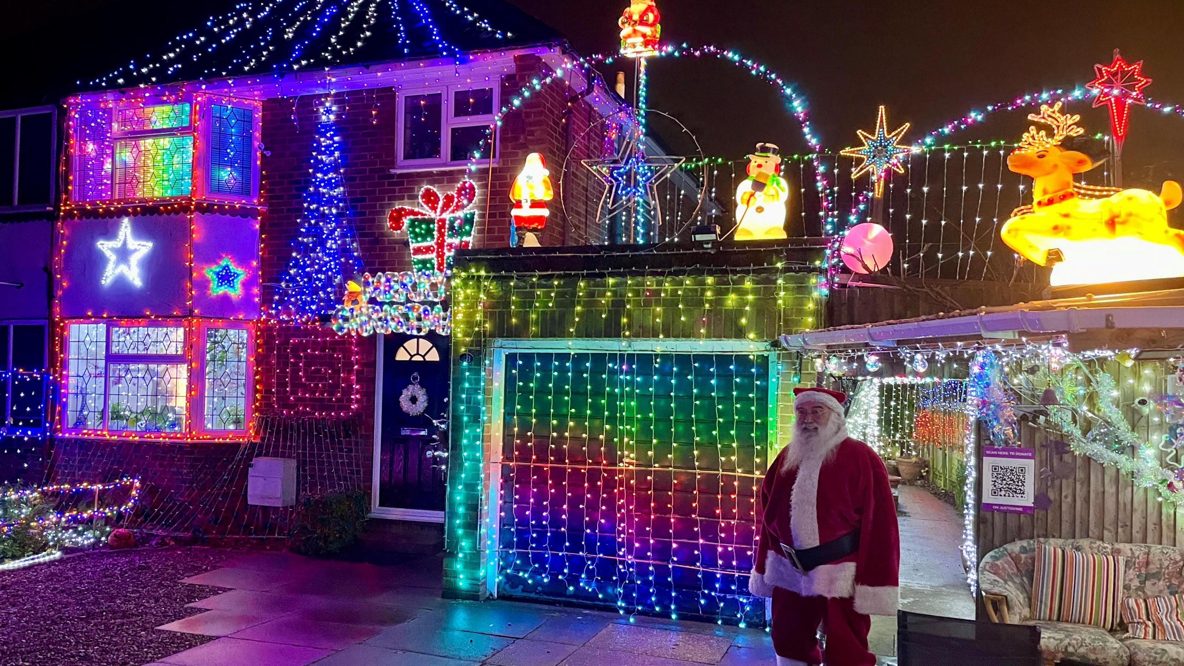 A wider shot of Mr O'Toole's 1950s-style semi-detached house covered in Christmas lights. Mr O'Toole is on the left dressed as Santa, standing at the entrance of a grotto.