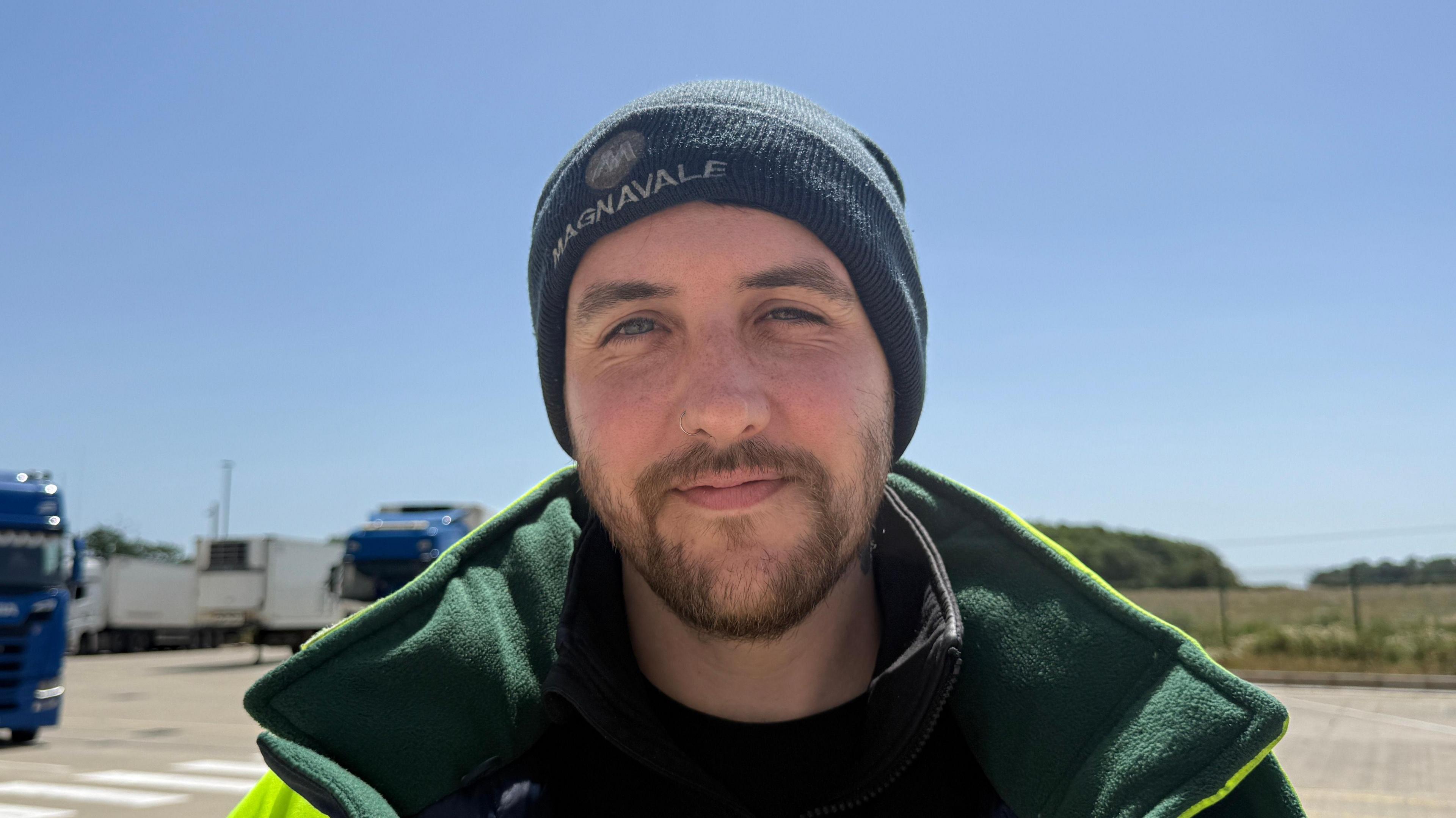 A man with a brown beard stands in a yard on a sunny day while wearing a green woolly hat and a yellow and green coat. HGVs can be seen behind him and the sky is blue.