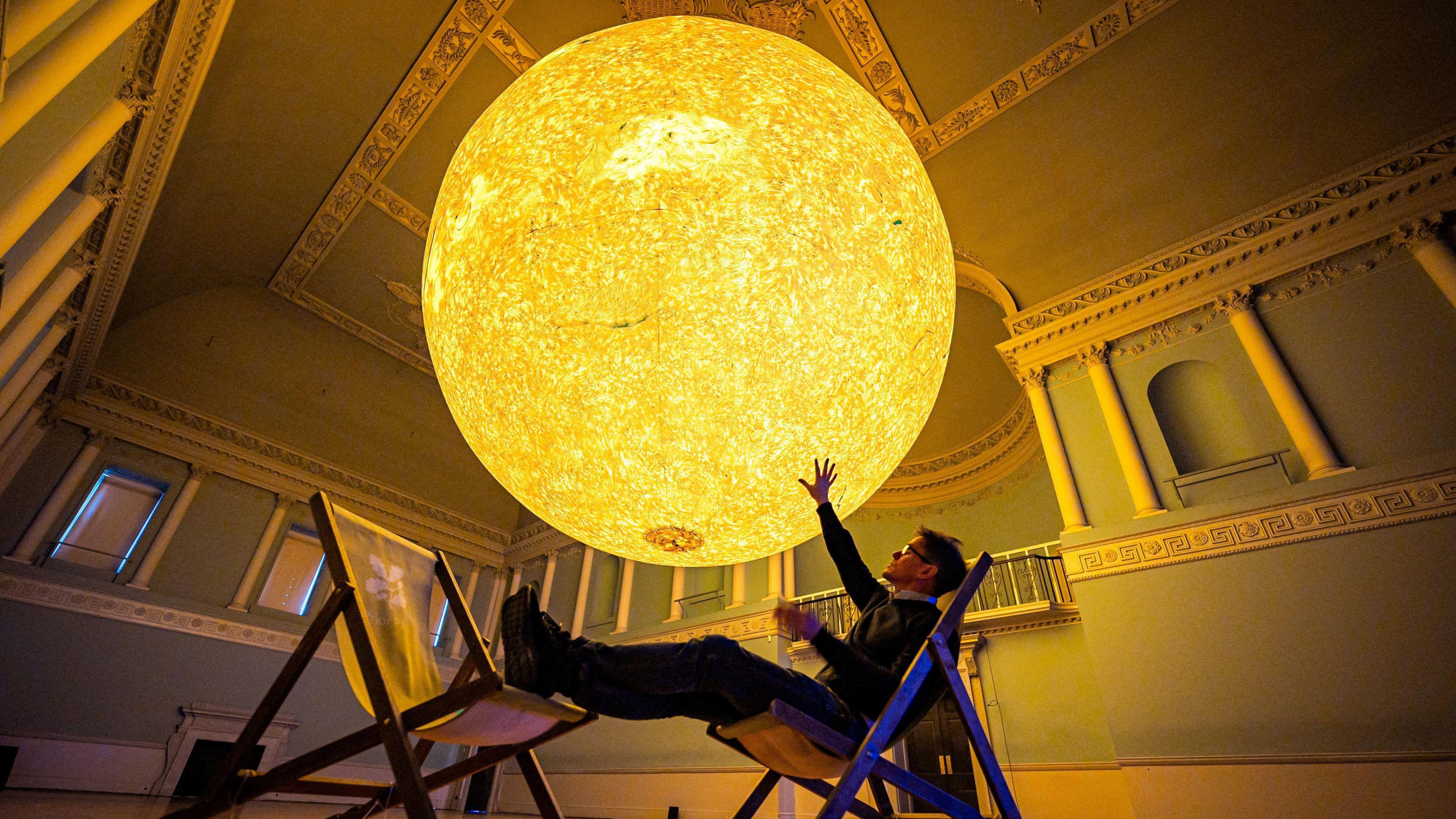 The artist Luke Jerram sits in a deckchair with his feet on another deckchair underneath a large glowing replica of the sun. The photograph is taken inside the Assembly Rooms in Bath. The artwork is called Helios