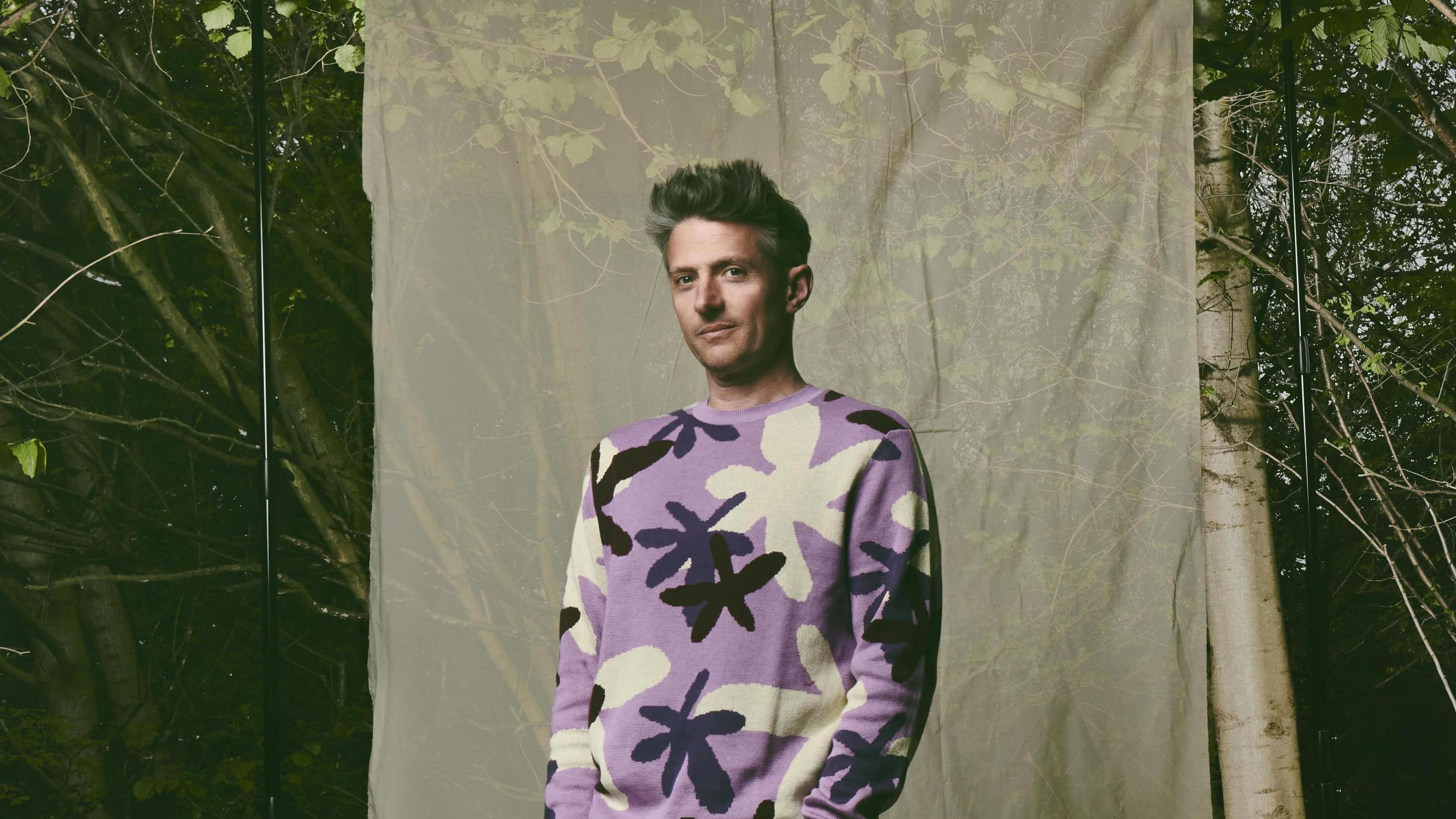 Stuart Goldsmith is standing in a forest in front of a sheet hanging from the trees. He is wearing a purple jumper with flowers and grey jeans.