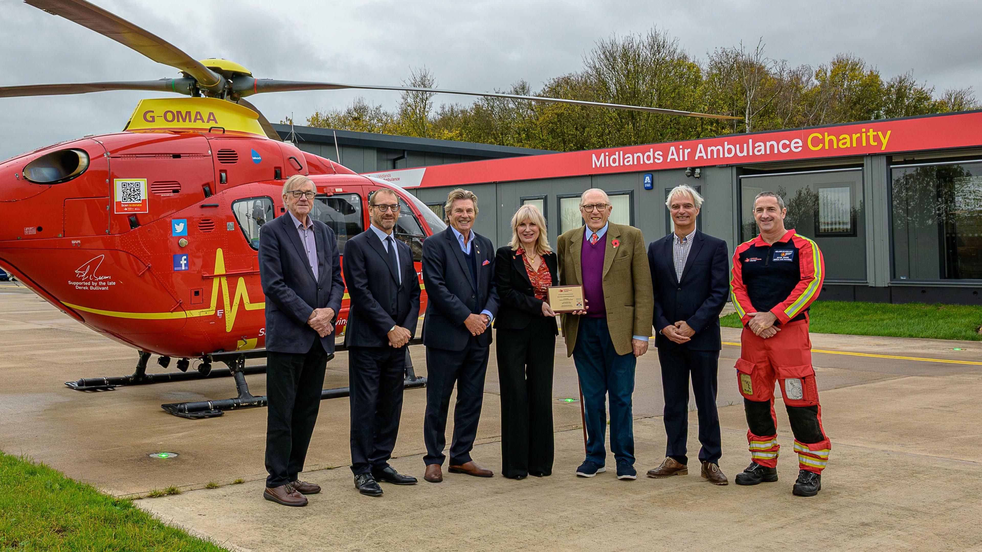 A group photo of seven people stand in front of a red and yellow air ambulance helicopter with a single storey building behind them with a sign reading "Midlands Air Ambulance Charity". 