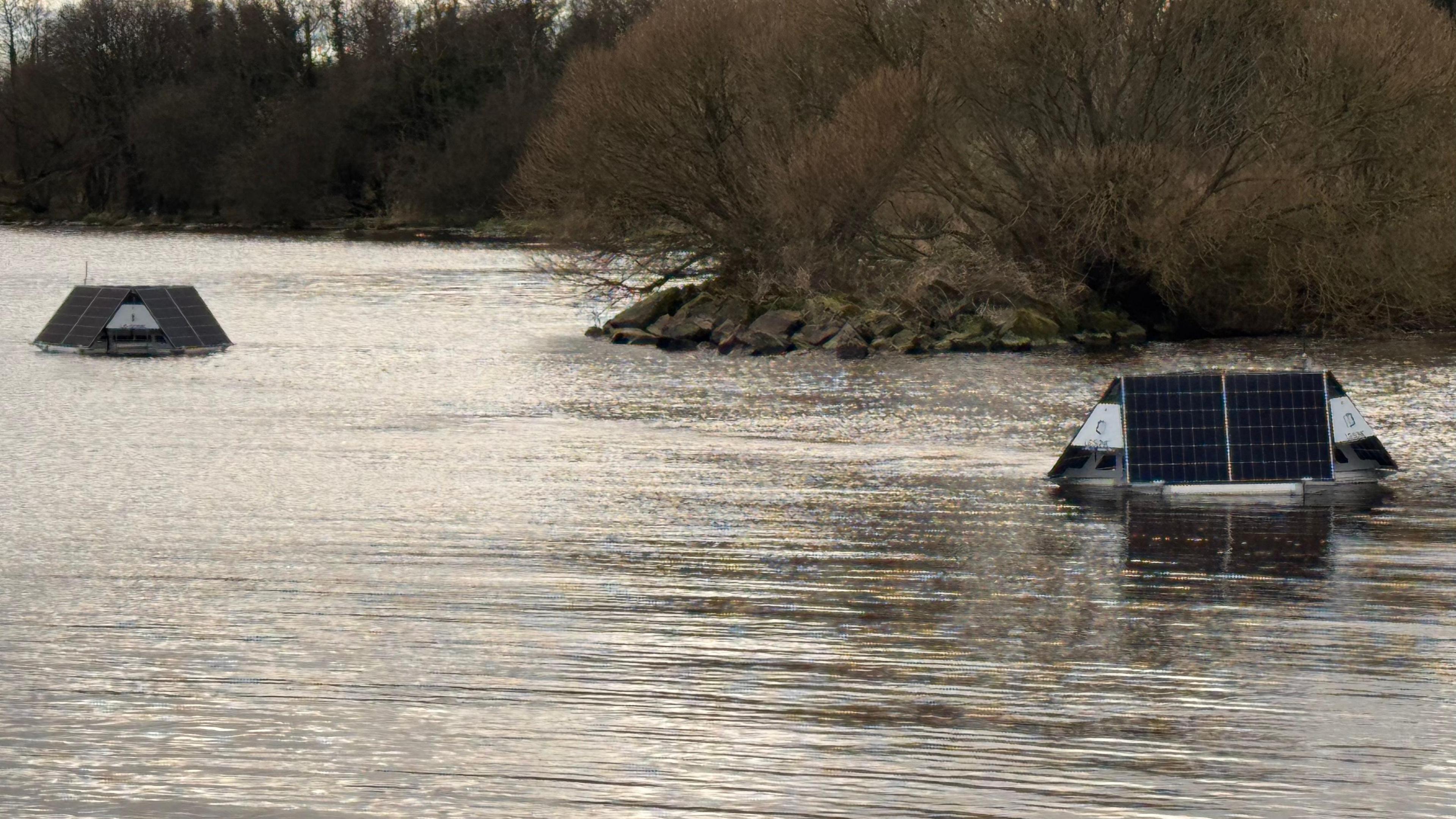Two floating bouys have small black solar devices on them. They float on a body of water on a cloudy day. Bare brown trees sit in the background.
