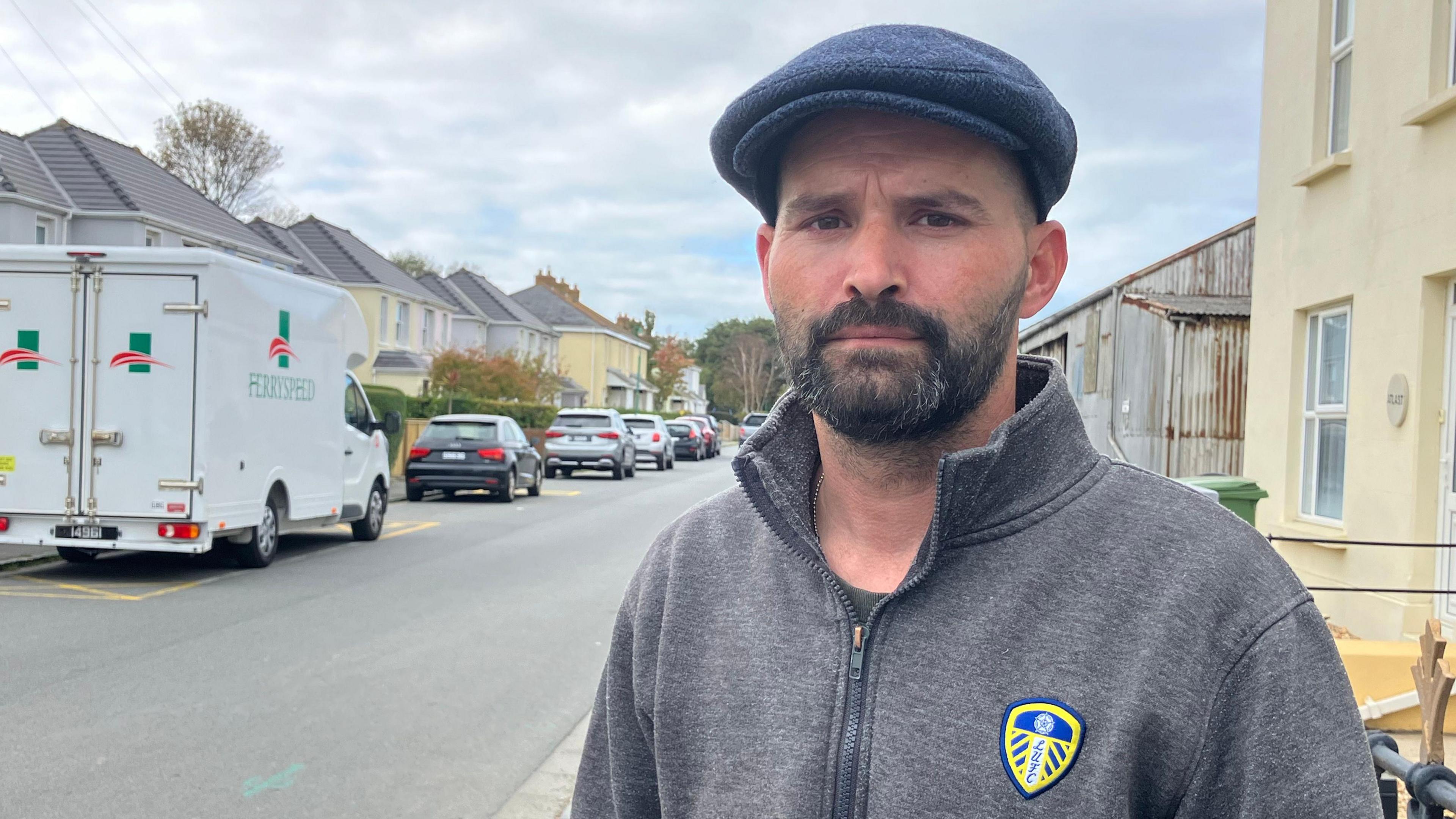 A man in a grey zip up hoodie. He is looking at the camera. He is wearing a blue cap and has a black beard. Behind him is a road. There are cars parked along it. The sky is grey.