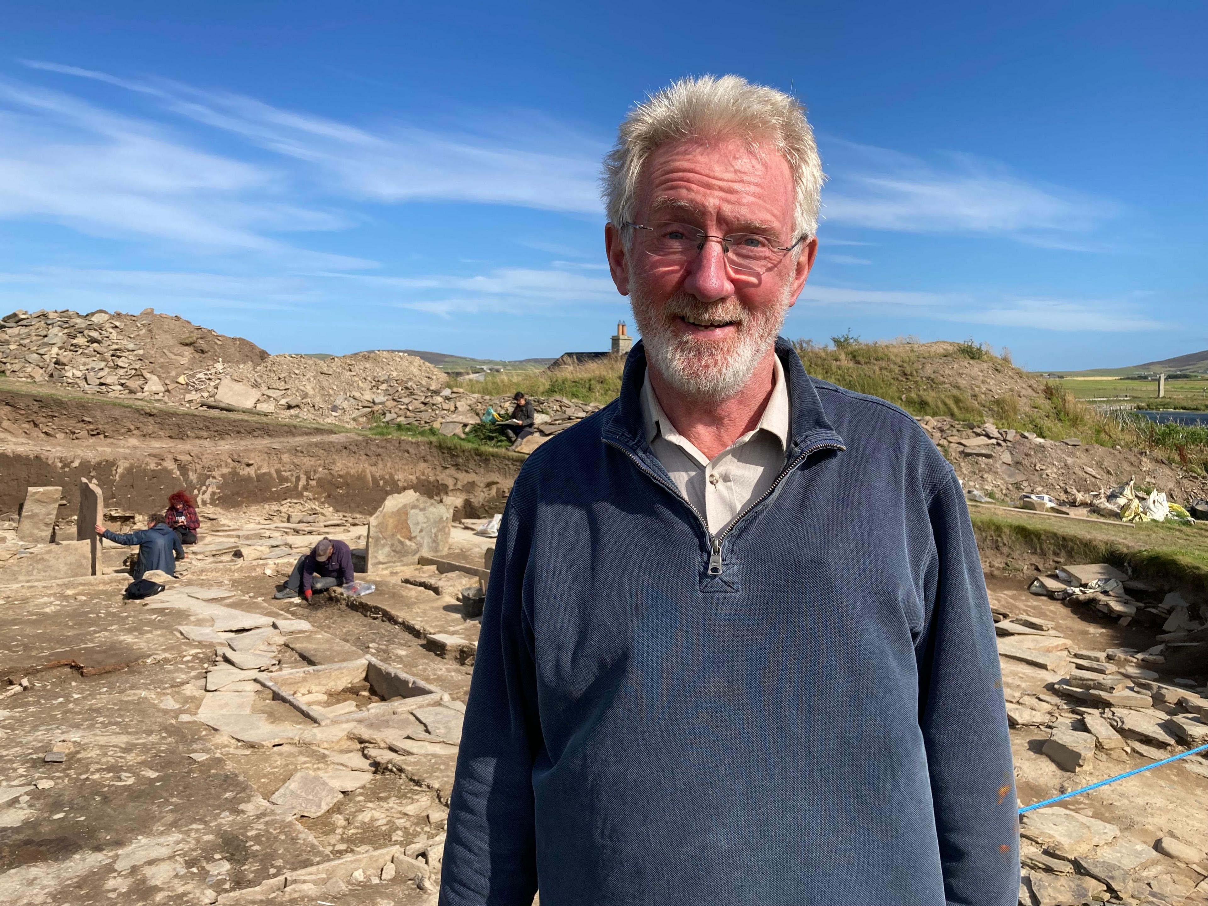 Archaelogist Nick Card standing by the side of the dig with people working behind him