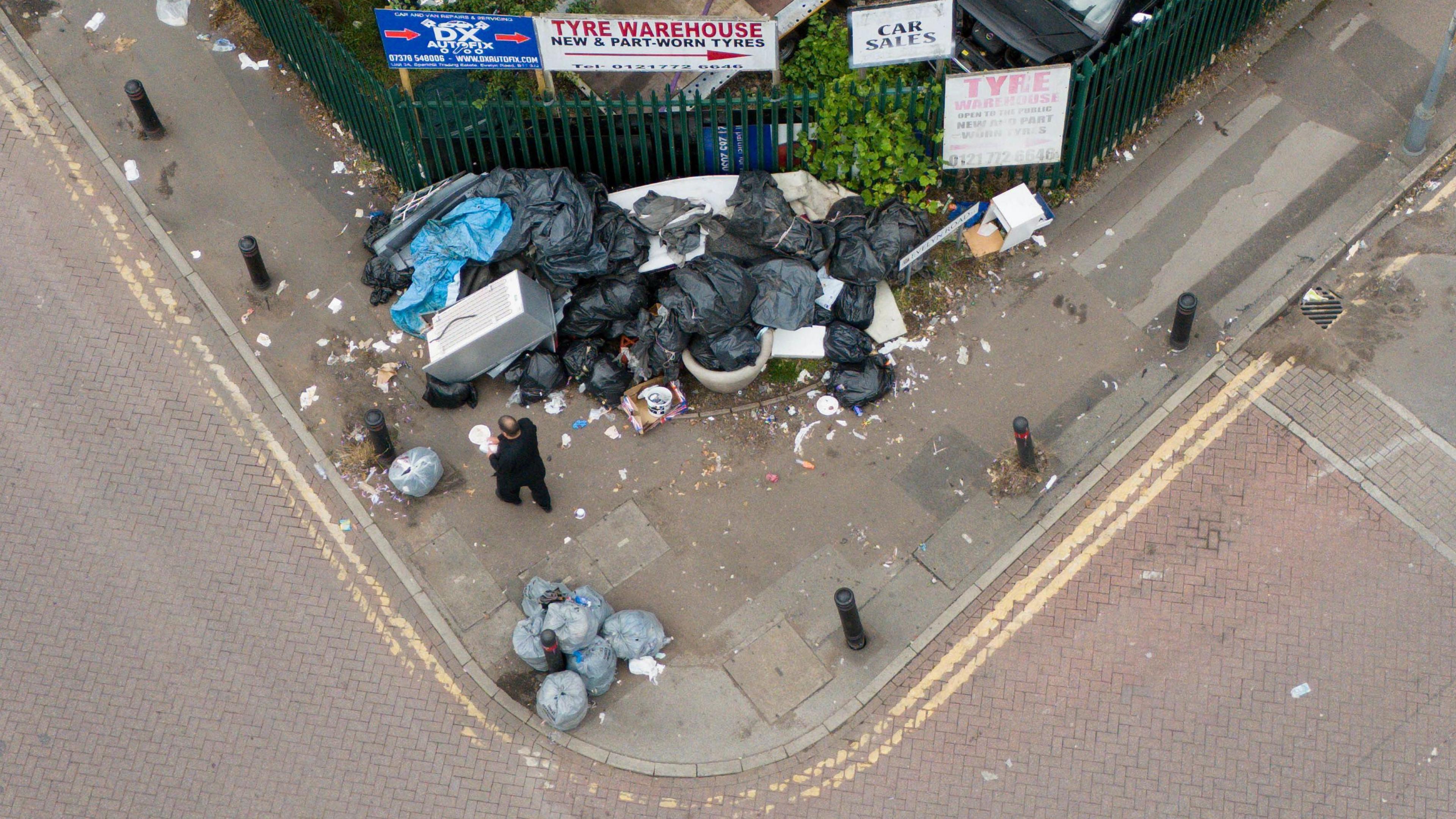 An aerial view of uncollected grey refuse bags piled by railing on a street corner, with a man walking past.