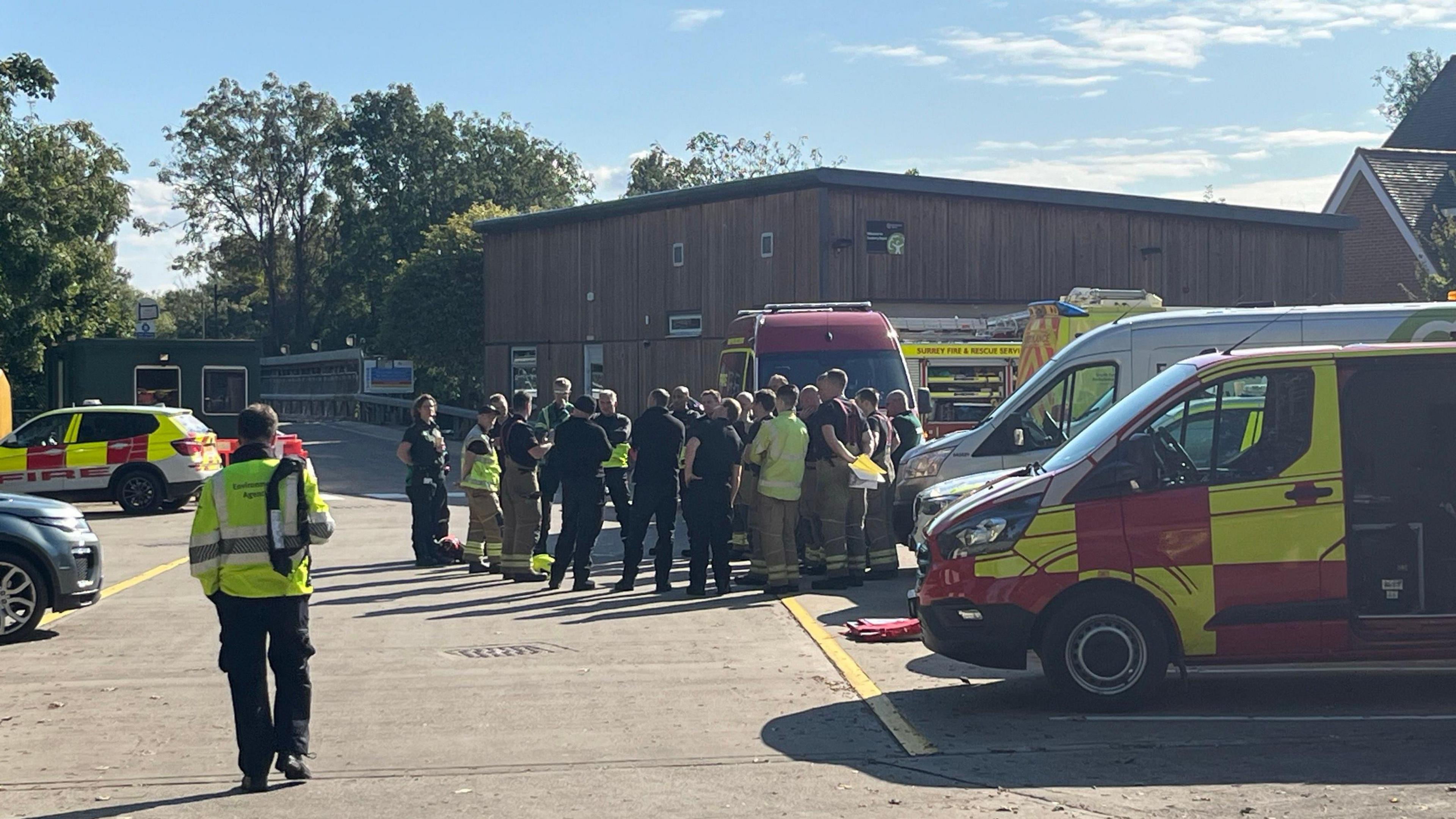 Emergency vehicles and workers are seen standing in a car park in front of a wooden building. Some are wearing hi-viz clothing and others in plain dark clothes.