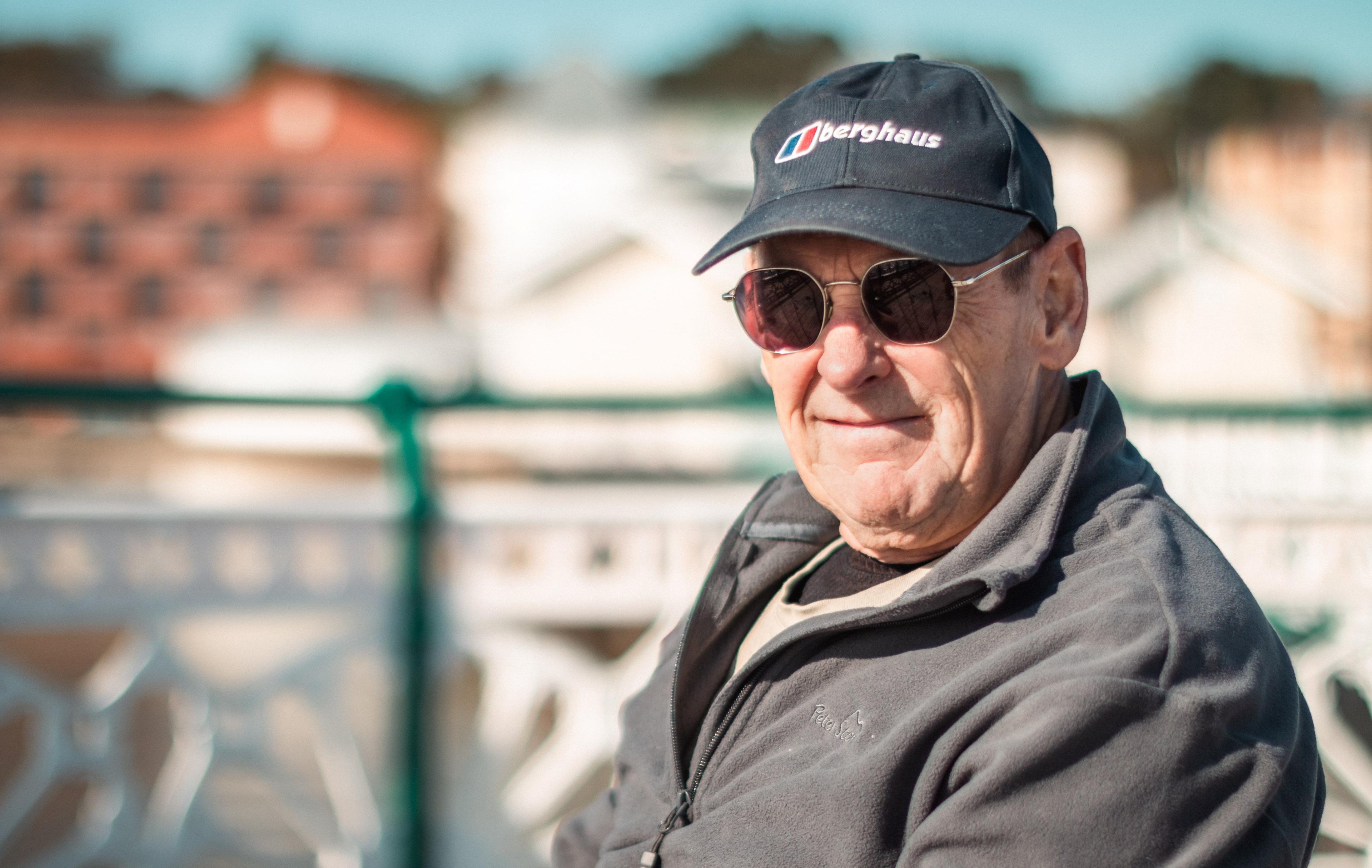 John Atkinson, who is sitting on Penarth pier, looking into the camera and wearing a grey baseball cap and glasses