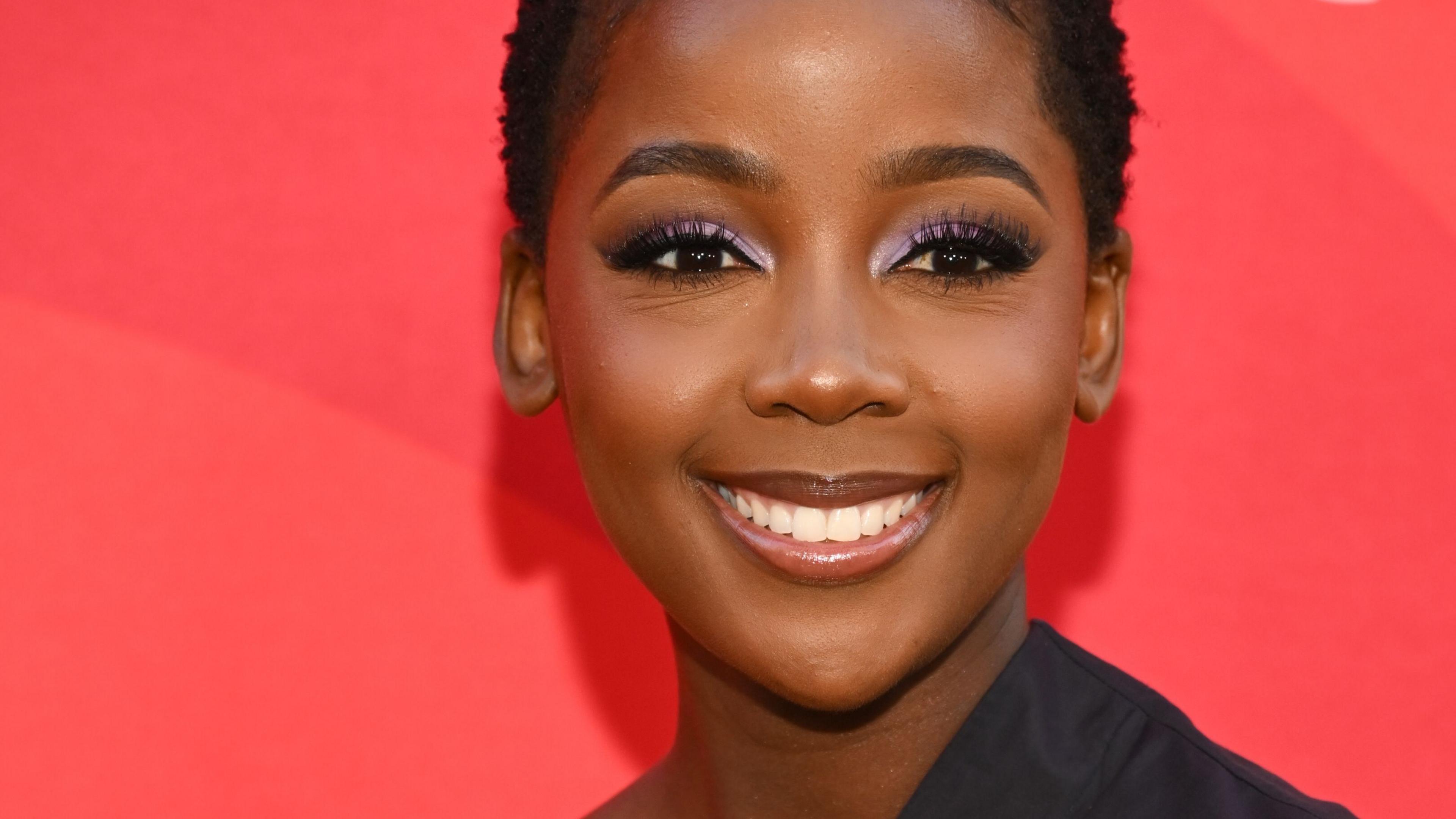 A young, smiling woman with cropped hair and a black outfit stands in front of a red background posing for photos.