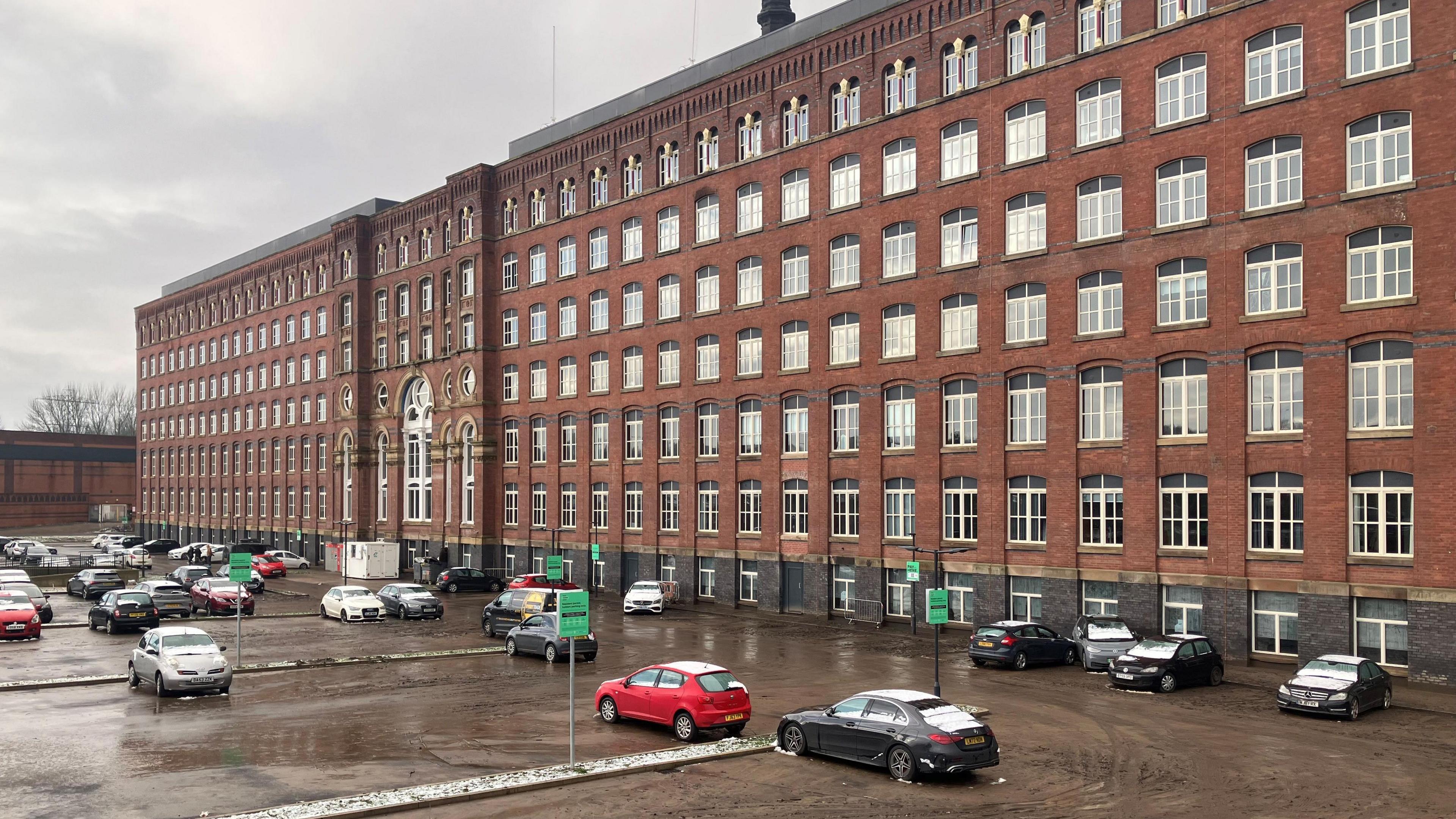 Meadow Mill in Stockport seen from the street, with mud covering the car park, where there are few snow-covered cars. 