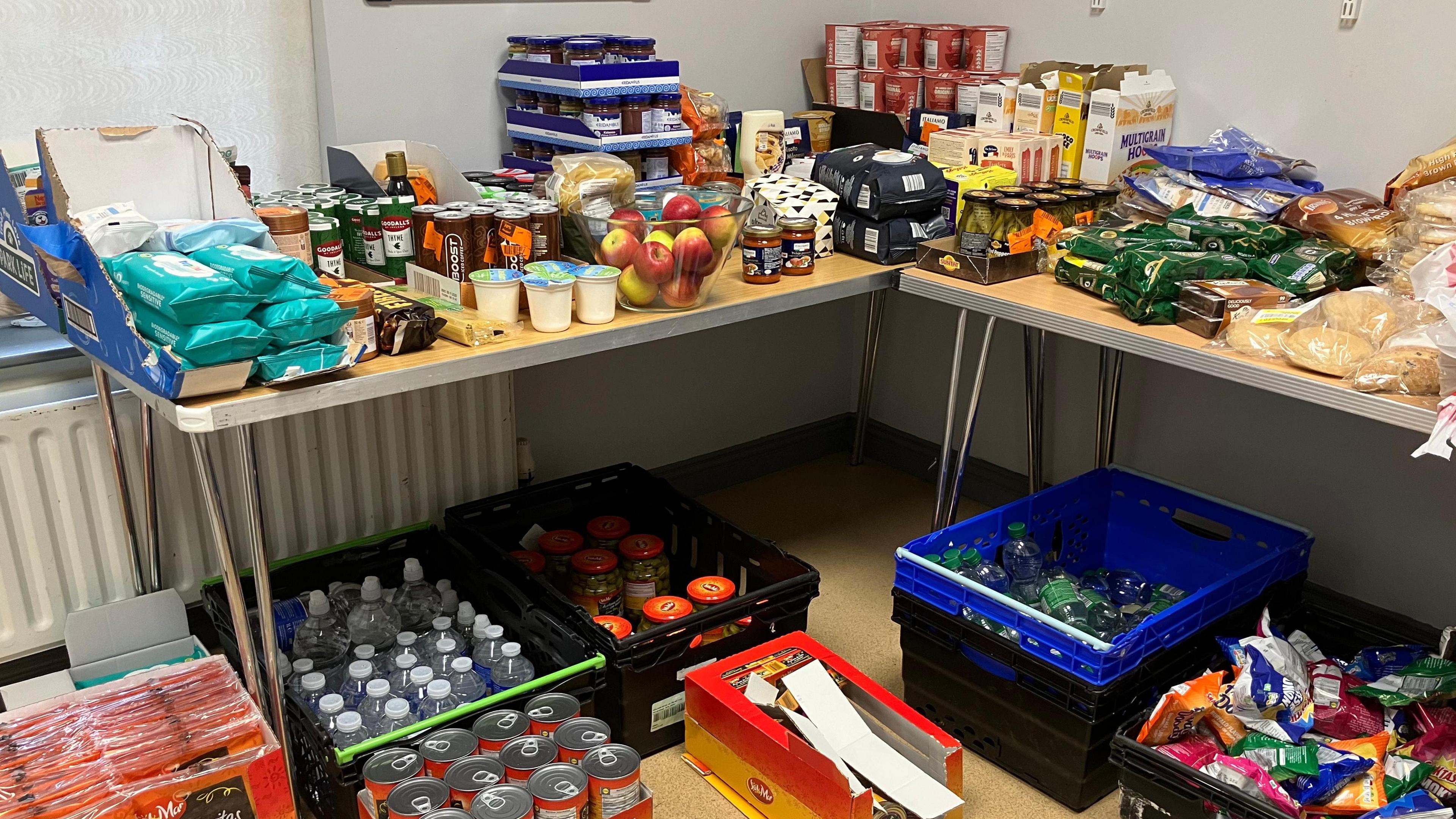 A corner of a room is filled with a colourful collection of cupboard items.  In the foreground there are boxes of bottled water and packets of crisps.  On the table sits a bowl of apples; loaves of many varieties of bread; cereals; jams ; pots of custard and ready made porridge in pots.