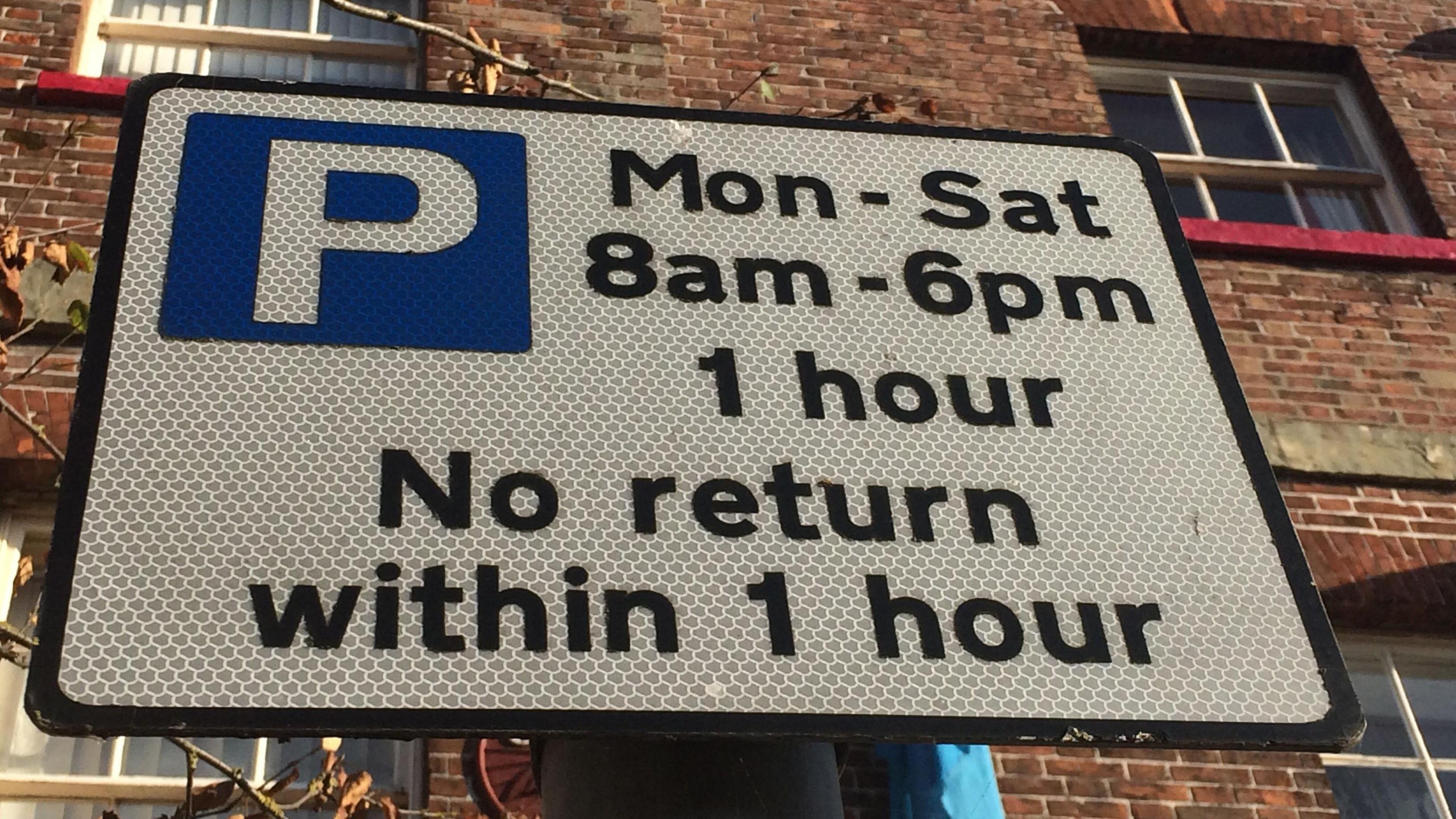 A generic view of a white on-street parking sign. It details when drivers are able to park on the road. A red brick building can be seen behind it.