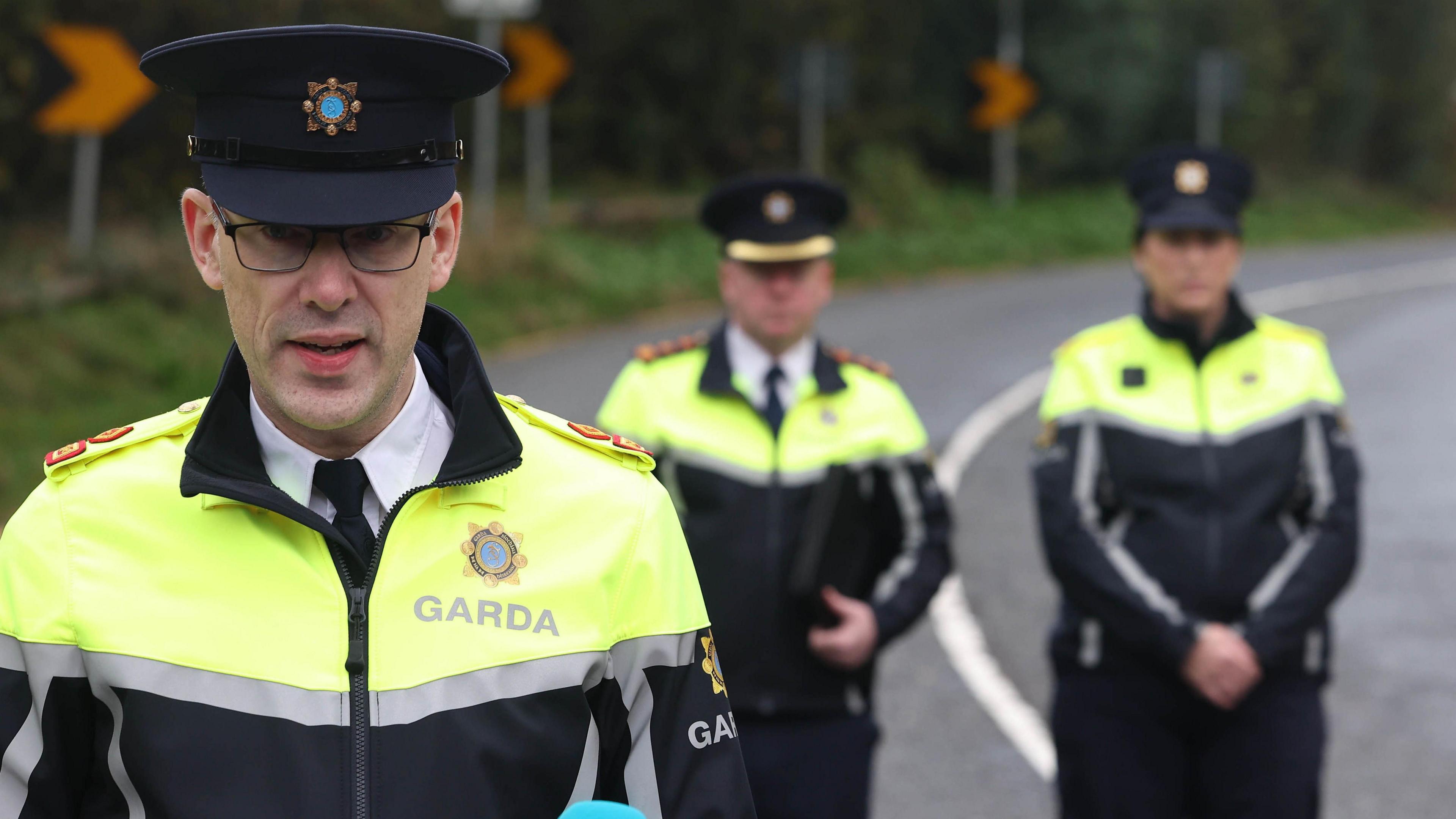 Supt Charlie Armstrong speaking to the media near the scene, there are two officers standing behind him. He is wearing a garda uniform with navy hat and galsses.