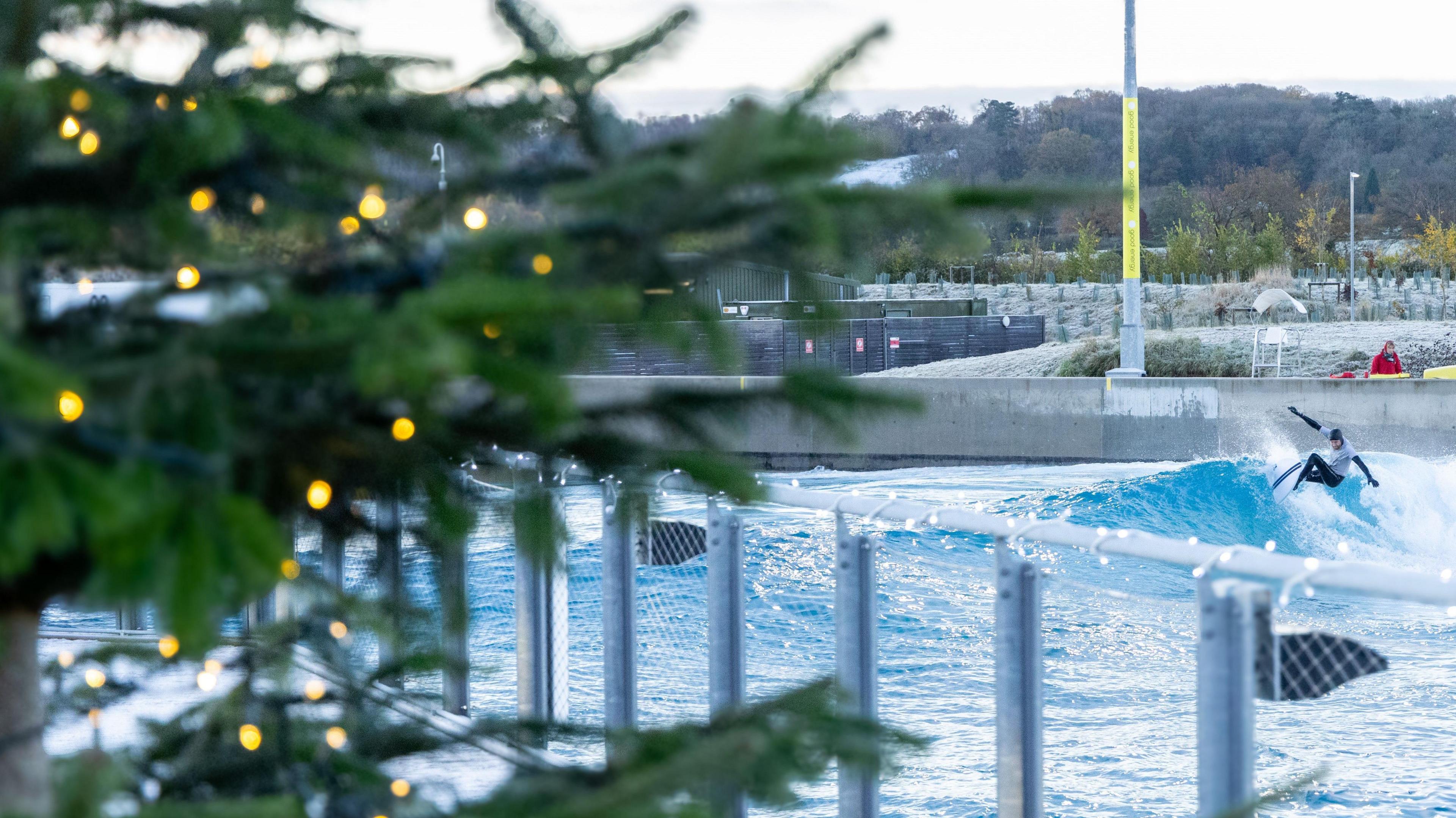 A Christmas tree with its lights on is seen slightly blurred in the foreground while in the background a man surfs at The Wave near Bristol. The ground in the background shows signs of frost and light snowfall