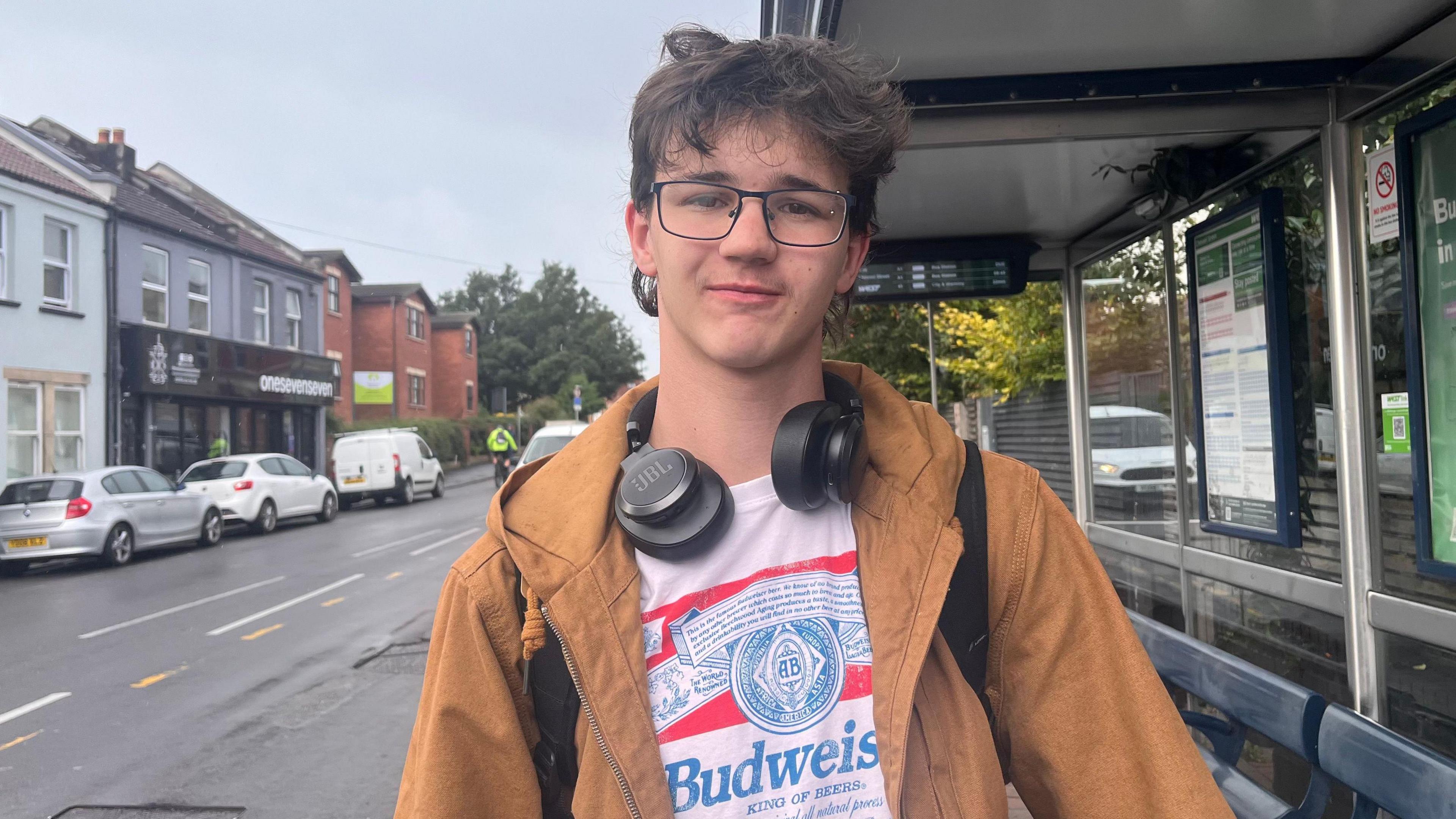 A young male stands under a bus stop, wearing a white t-shirt and caramel coat, he wears glasses and a pair of headphones hang around his neck.