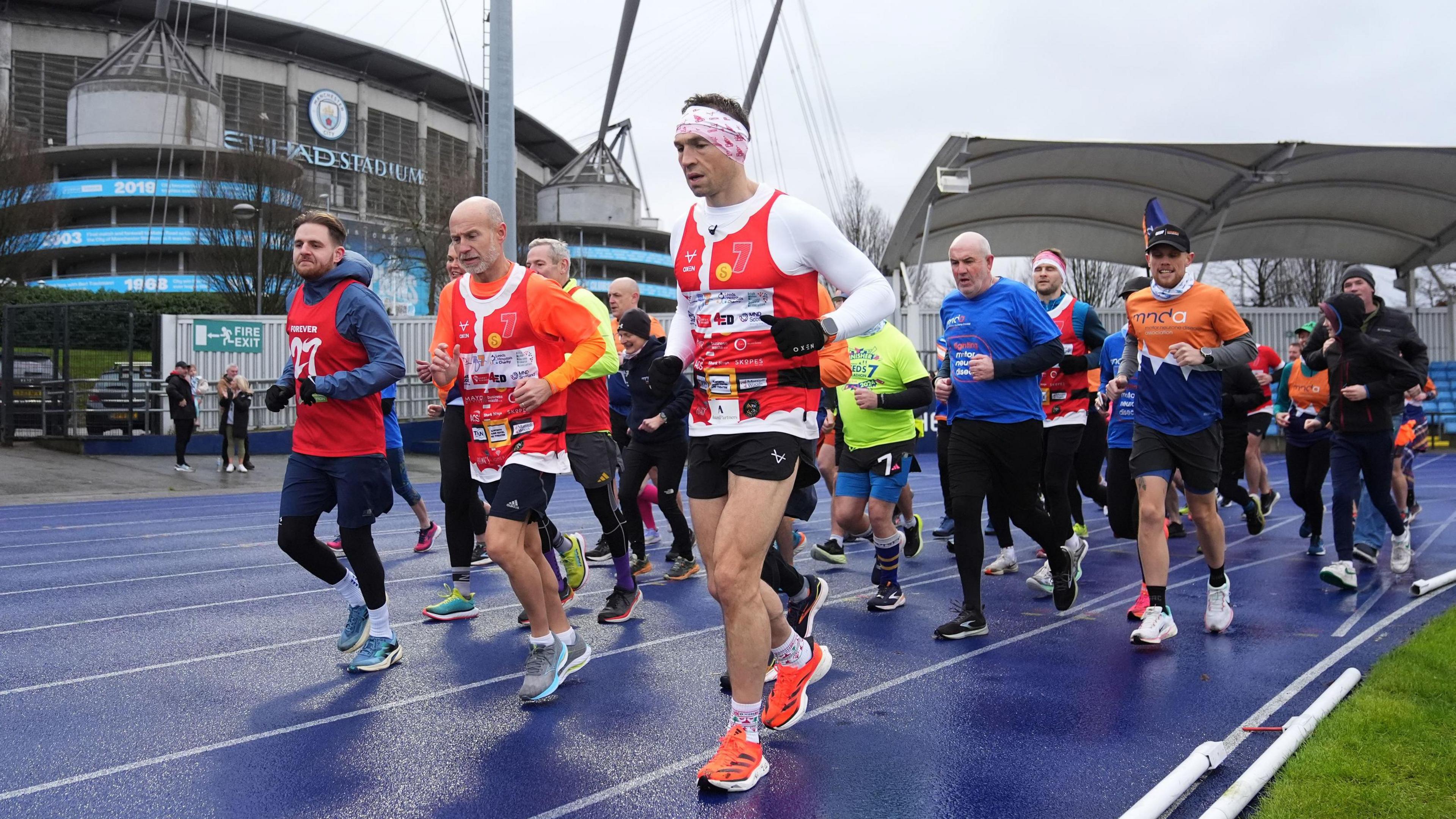 Sinfield accompanied by a large group of people running on the blue athletic track next to Manchester City's stadium