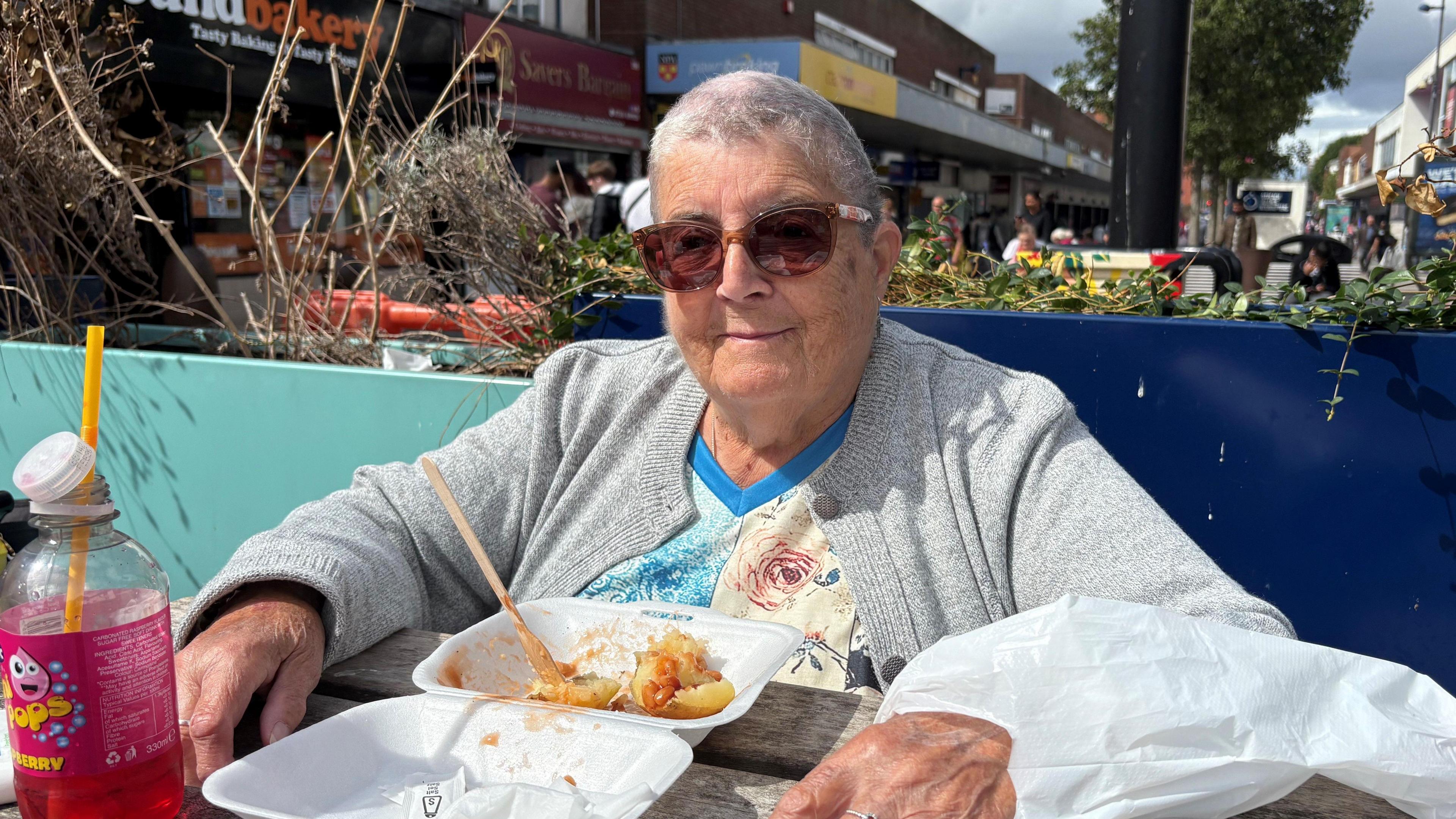A woman with short grey hair sits at a bench on the street wearing sunglasses and a grey cardigan having chips.