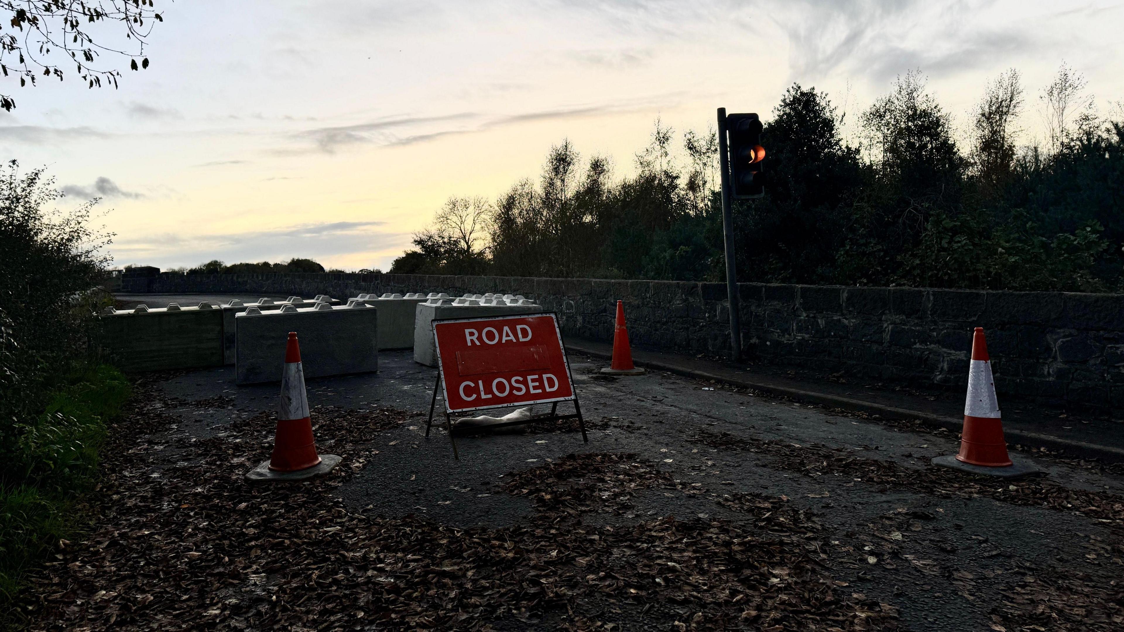 A road closed sign approaching Kilrea Bridge.  Traffic cones and large blocks have been placed across the road.  A single traffic light shows an amber signal.