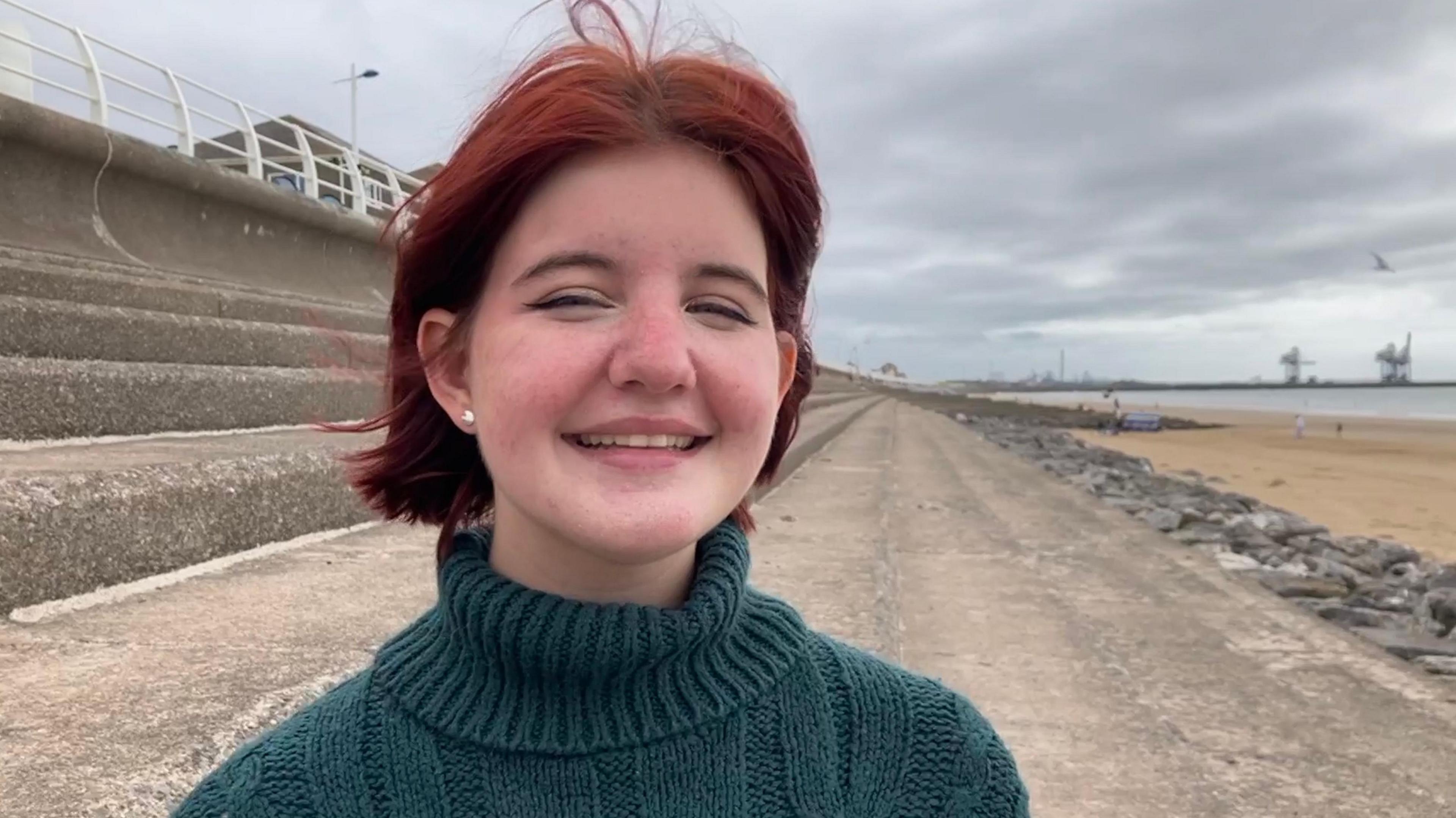 A young woman with short, red hair is looking towards the camera and smiling. She's wearing a turtle-neck, green jumper and an earing is visible on the left side. She's sitting on concrete steps which lead to the beach and in the background there are some large cranes and the steelworks.