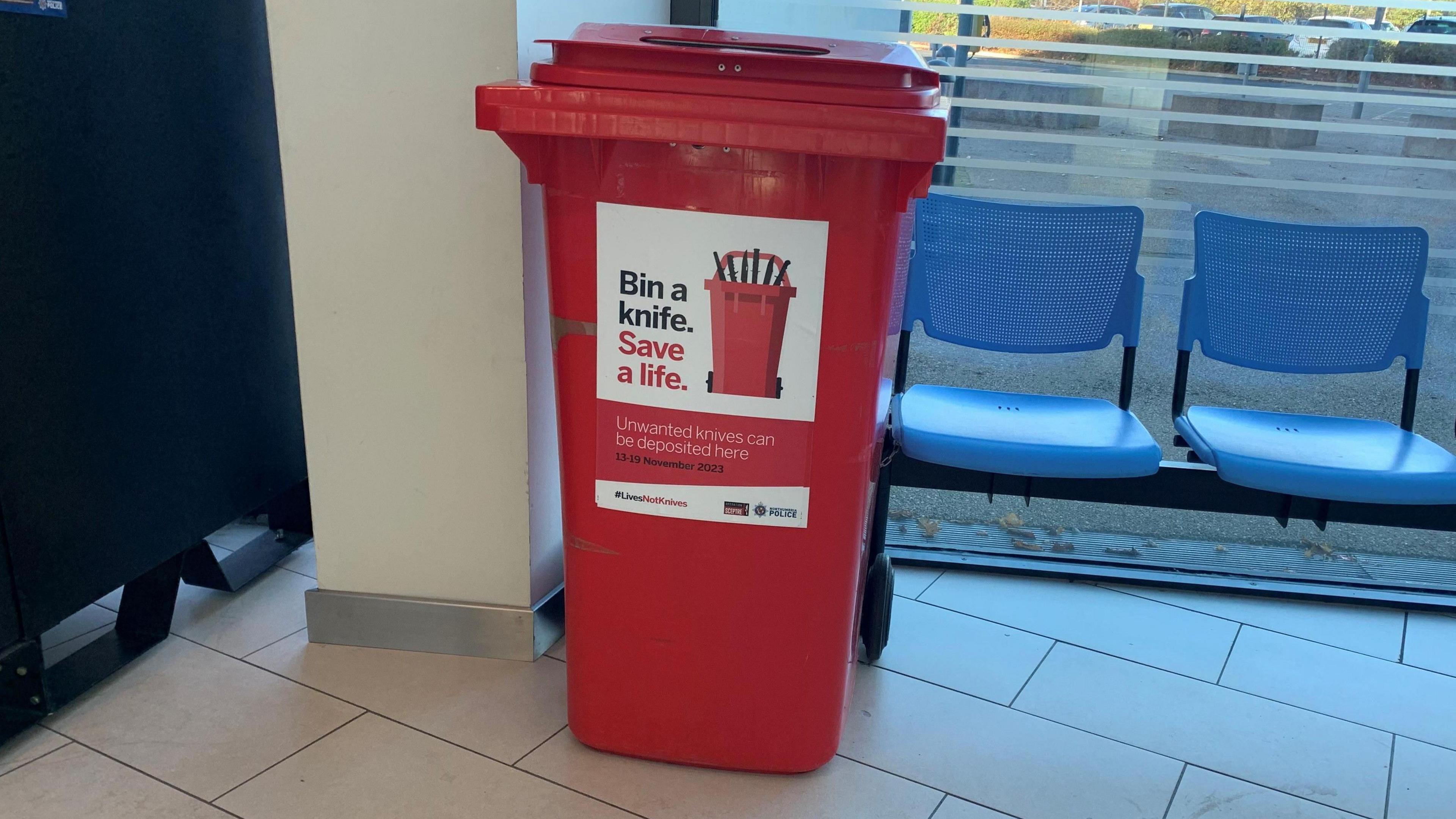 A red wheelie bin with a sign on it reading: "Bin a knife. Save a life."