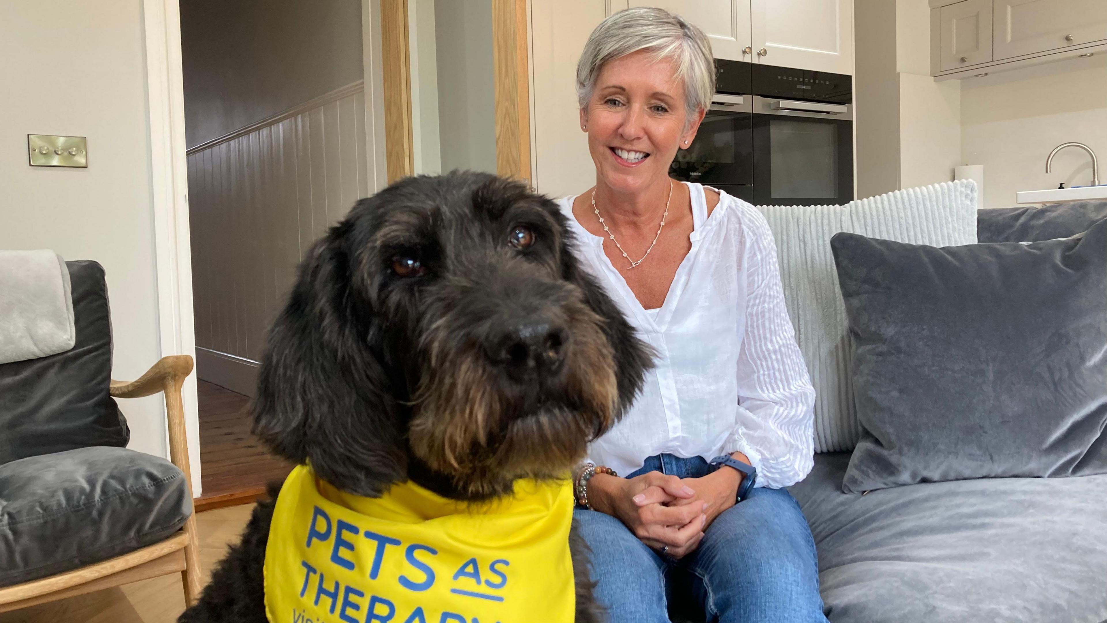 Poppy the labradoodle is sat in the foreground wearing a yellow neckerchief, which has 'Pets as therapy' printed on it in blue letters. She has brown eyes, long floppy ears and her hair is a slightly lighter shade of brown around her nose and mouth. Poppy's owner, Lisa, is sat on a light grey couch with her hands clasped in her lap, smiling directly at Poppy. Lisa wears a white blouse, blue jeans and has short light hair.