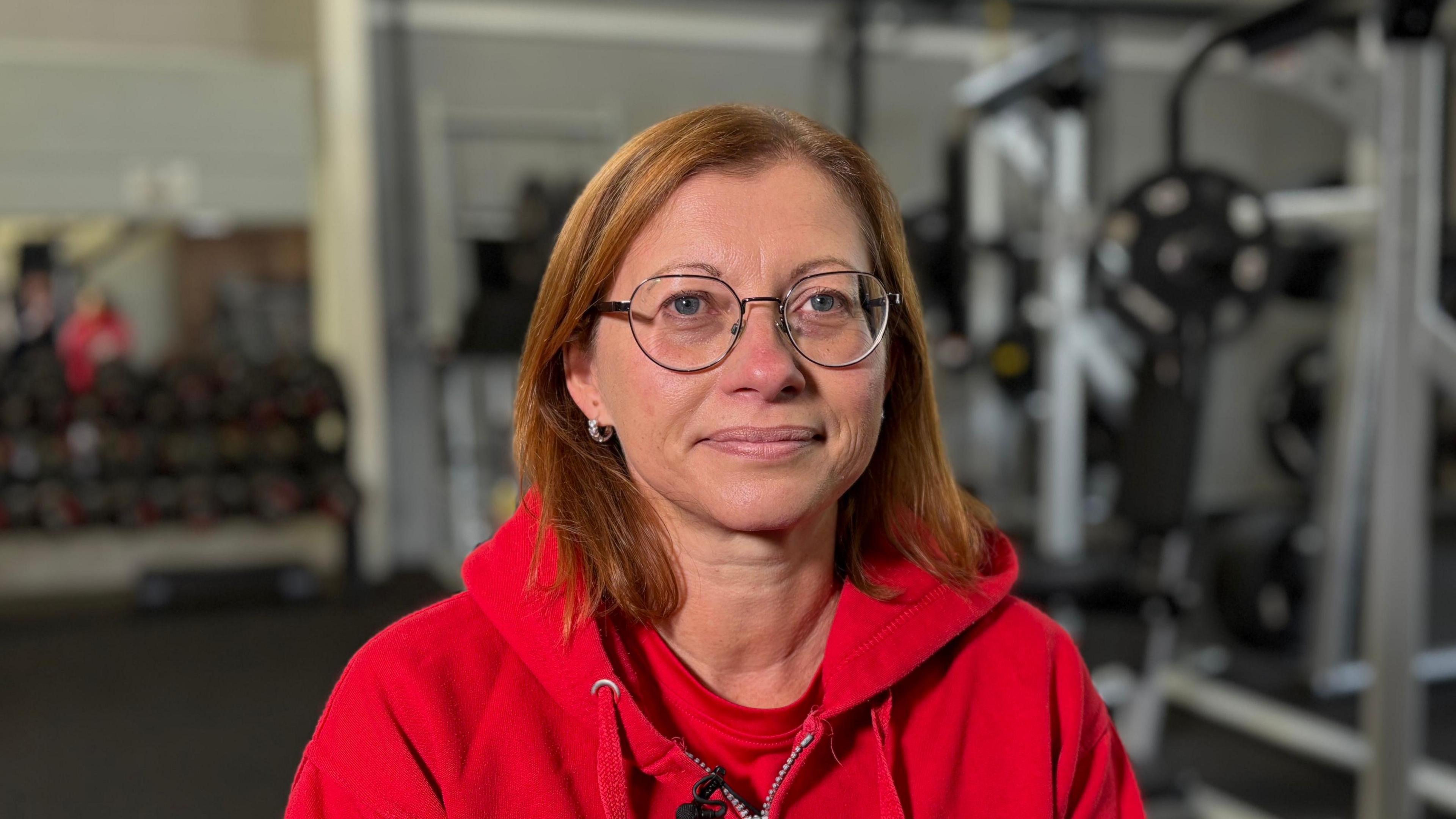 Mel Stollery looking directly down the camera. She is seated inside a gym area and is wearing a bright red hooded top. She is smiling slightly, and is wearing round-rimmed metal glasses. Her hair is a deep orange colour and falls to shoulder length. Where she has pulled it back over one ear, a small shining hooped earring can be seen. The background of the image is blurred, but gym equipment and a rack of weights can just be made out behind Mel.