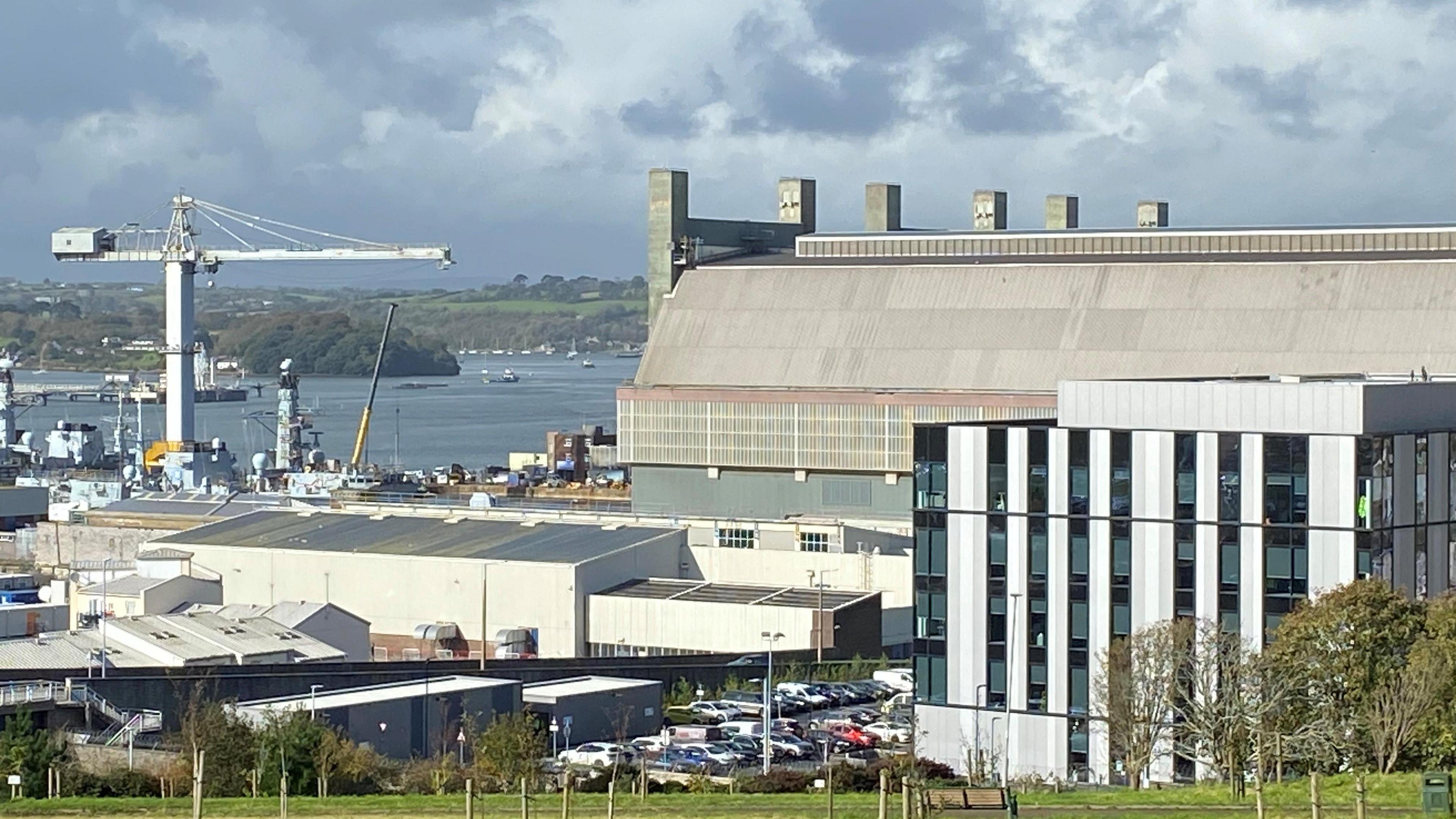 A general view of of the dockyard. Large buildings and a crane are visible. Water and boats are in the distance. The sky is grey and cloudy.