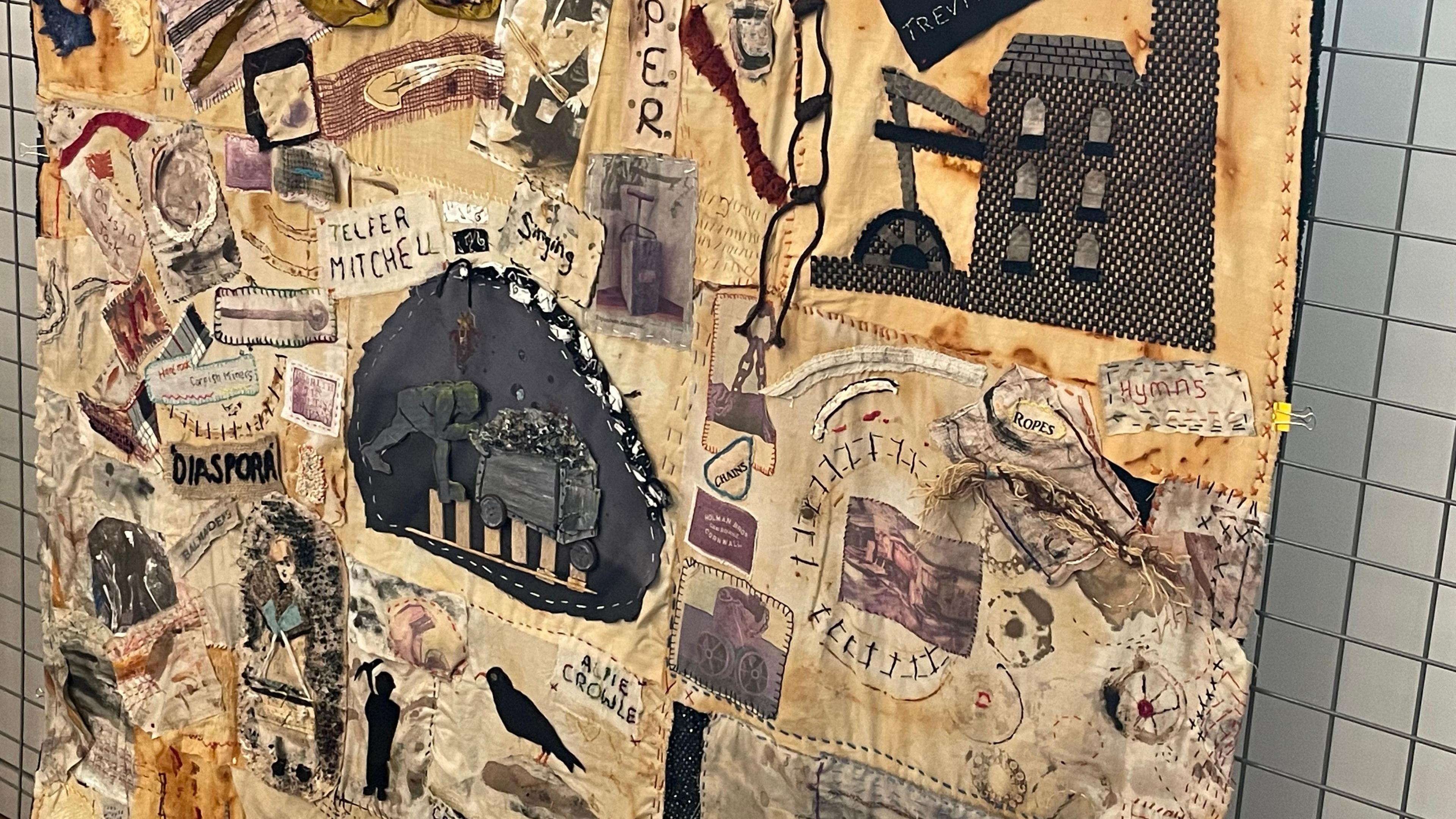 A photo of the wall hanging. It shows different bits of patchwork which relates to mining, including a black mining building on the left of the mural, and a man pushing coal down a track on the right.